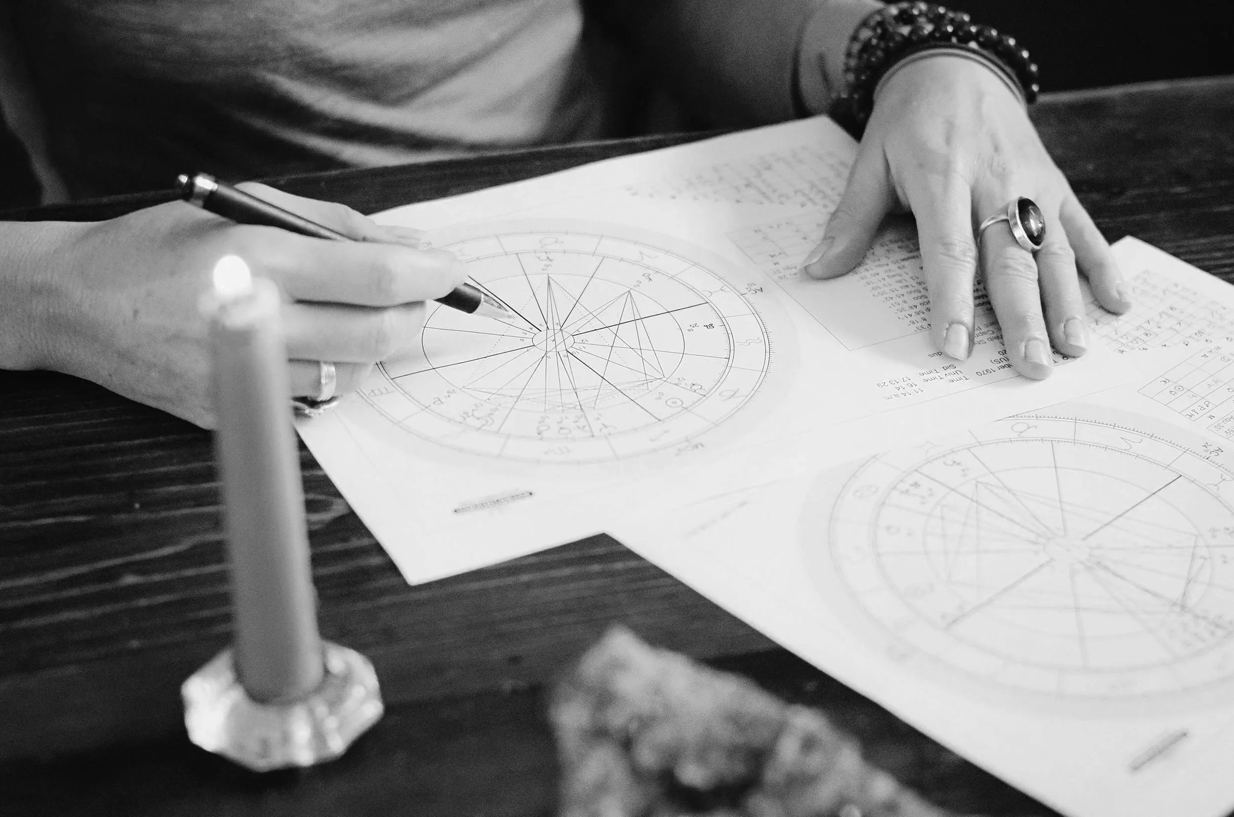 A person is working on detailed astrological or astronomical charts with a pen, while also reviewing handwritten notes on a desk. The person is wearing rings and bracelets.