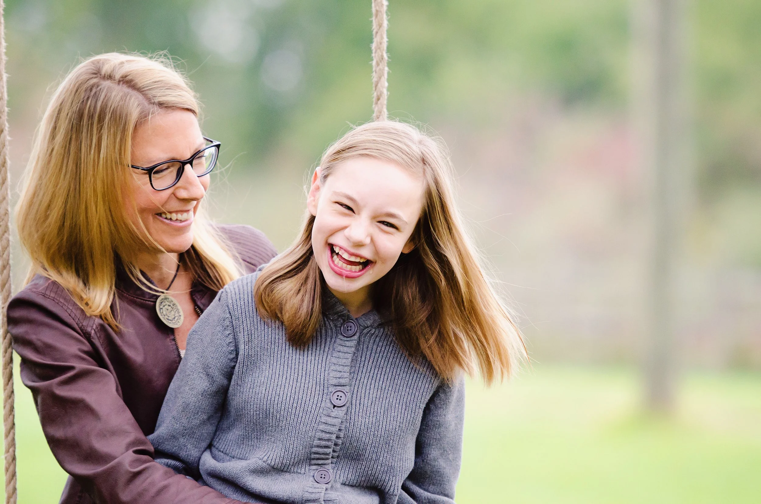 A woman with blonde hair, glasses, and a maroon jacket smiling and sitting on a swing with a young girl with light brown hair and a grey cardigan, both laughing outdoors.