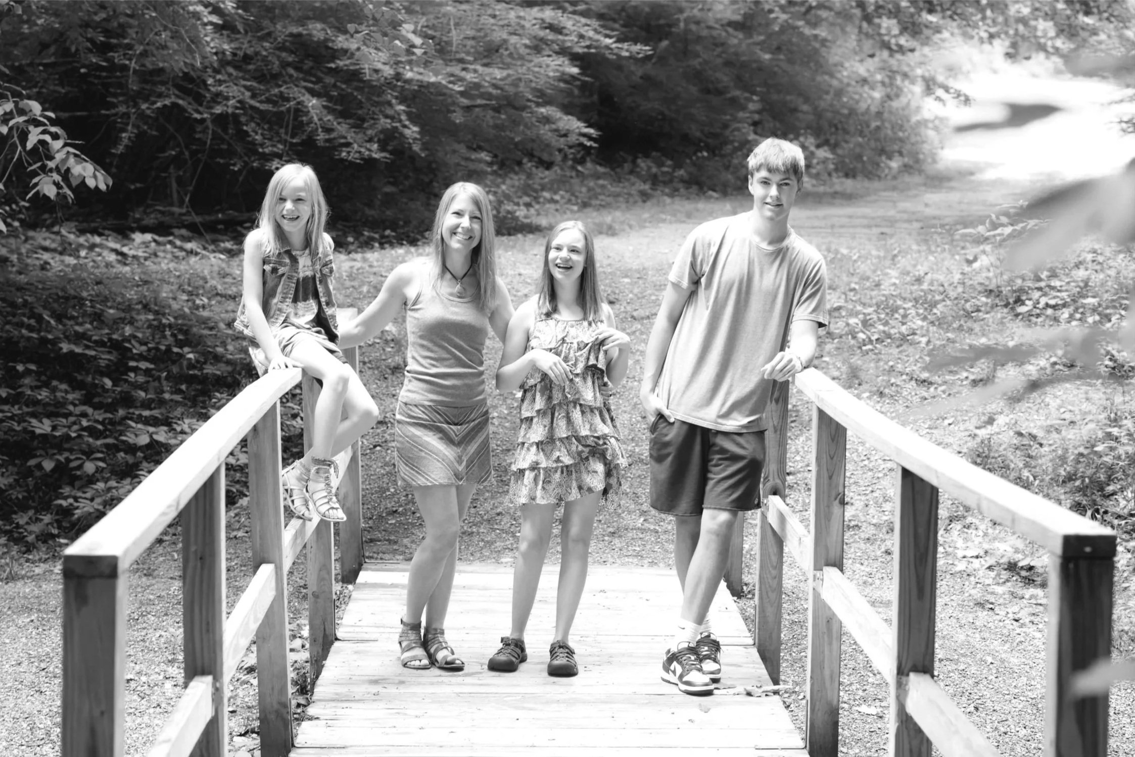 Four kids standing on a small wooden bridge in a wooded area, smiling and enjoying their time outdoors.