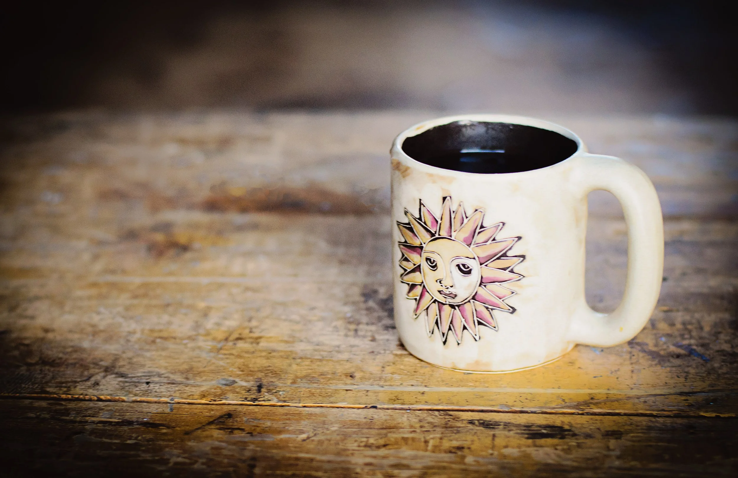 Cream-colored coffee mug with a sun and moon face design, filled with black coffee, placed on a rustic wooden surface.