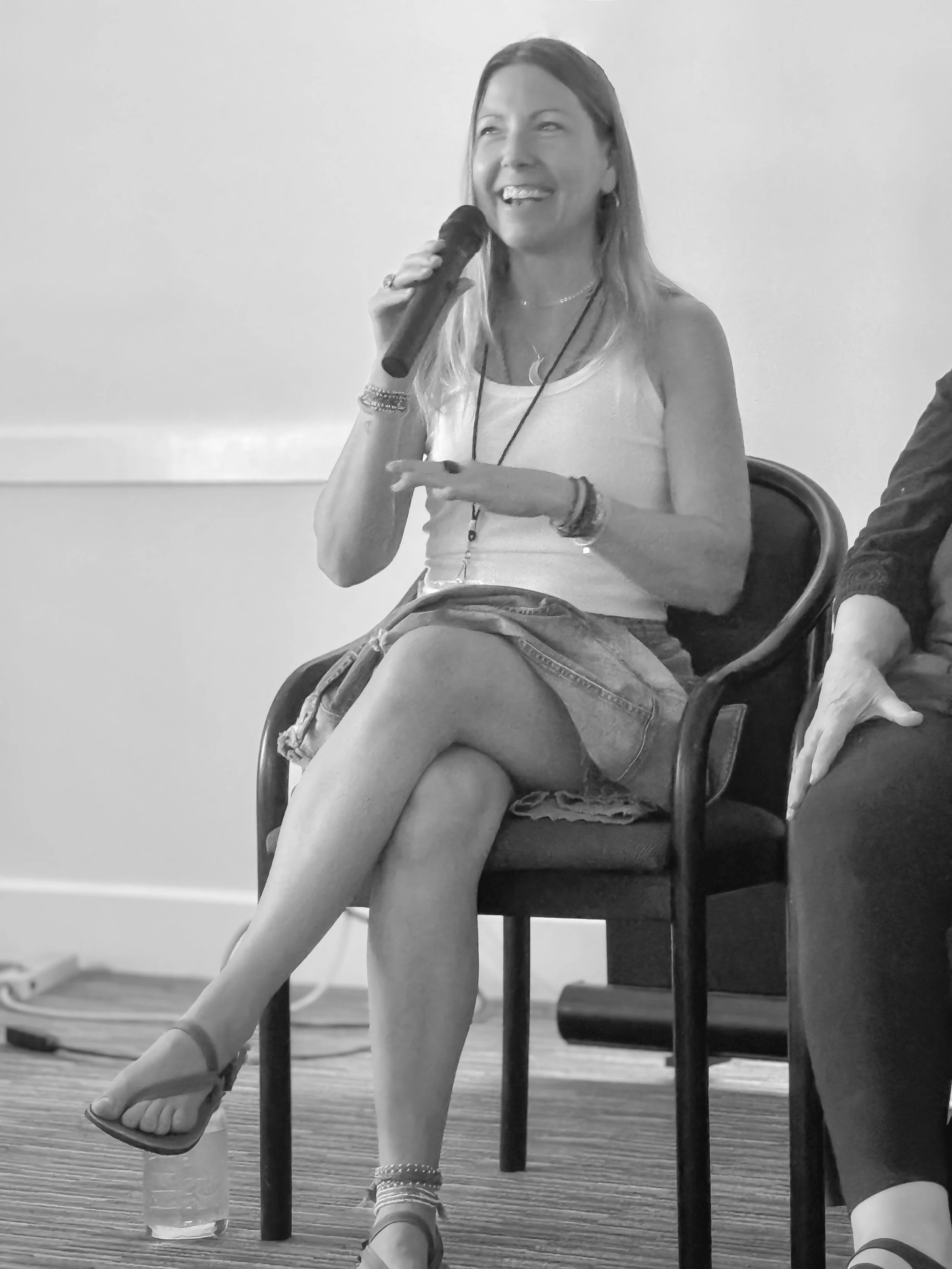 Parenting Astrologer, Tara Vogel, a woman sitting on a chair, holding a microphone, smiling, wearing a sleeveless top, shorts, and sandals.