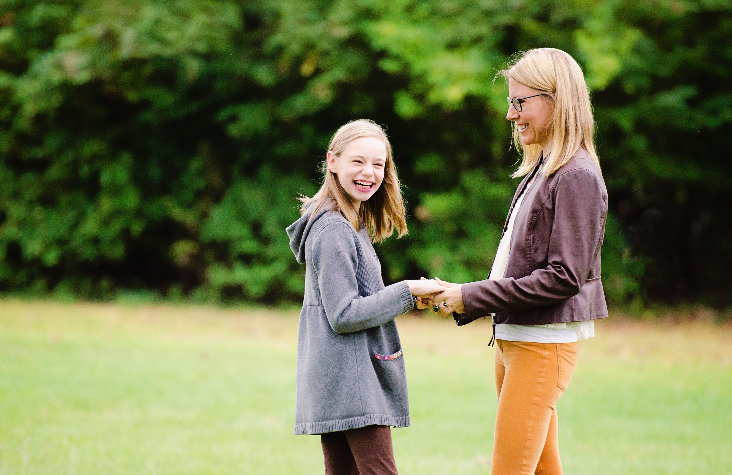 A woman and girl laughing and holding hands outdoors in a grassy area with trees in the background.