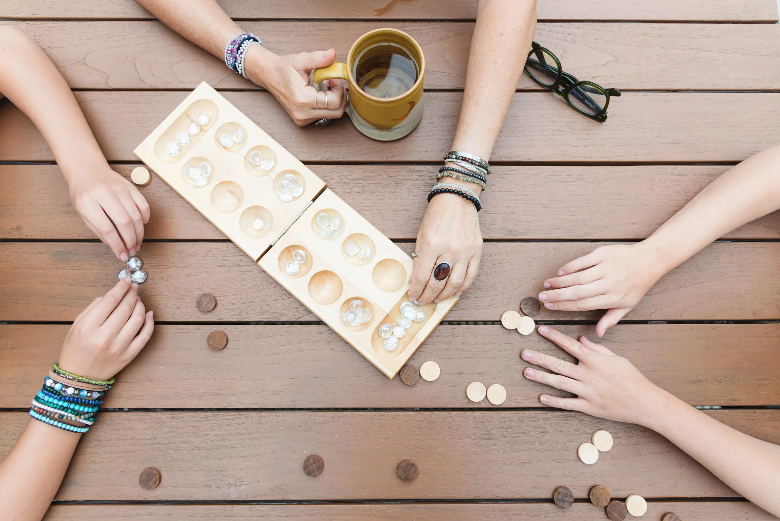 Three people playing a tabletop game with wooden pieces, a mug of coffee, and glasses on a wooden table.
