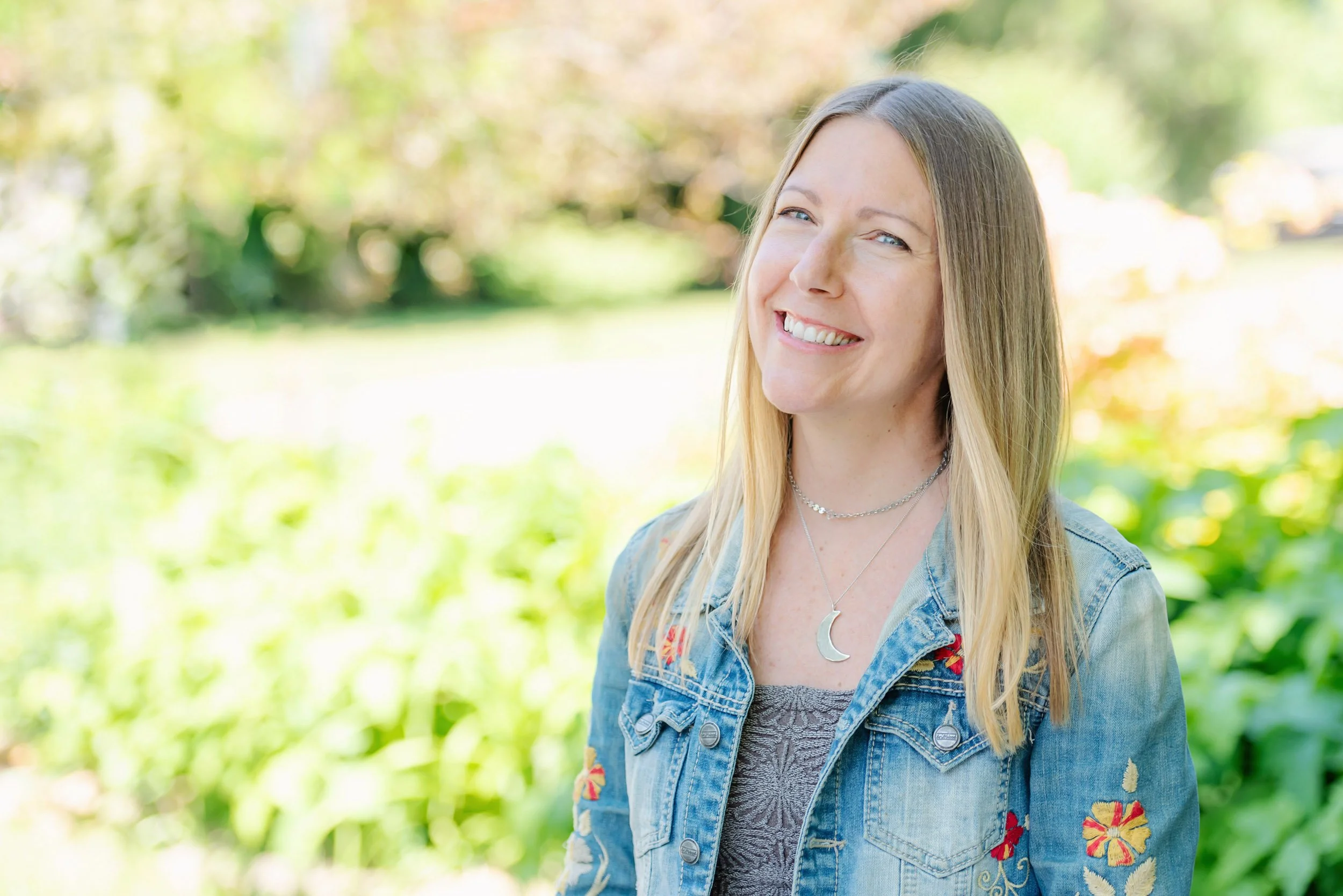 Parenting Astrologer, Tara Vogel, a smiling woman with long blonde hair, standing outdoors in a garden or park during daytime. She is wearing a denim jacket with floral embroidery and a moon necklace.