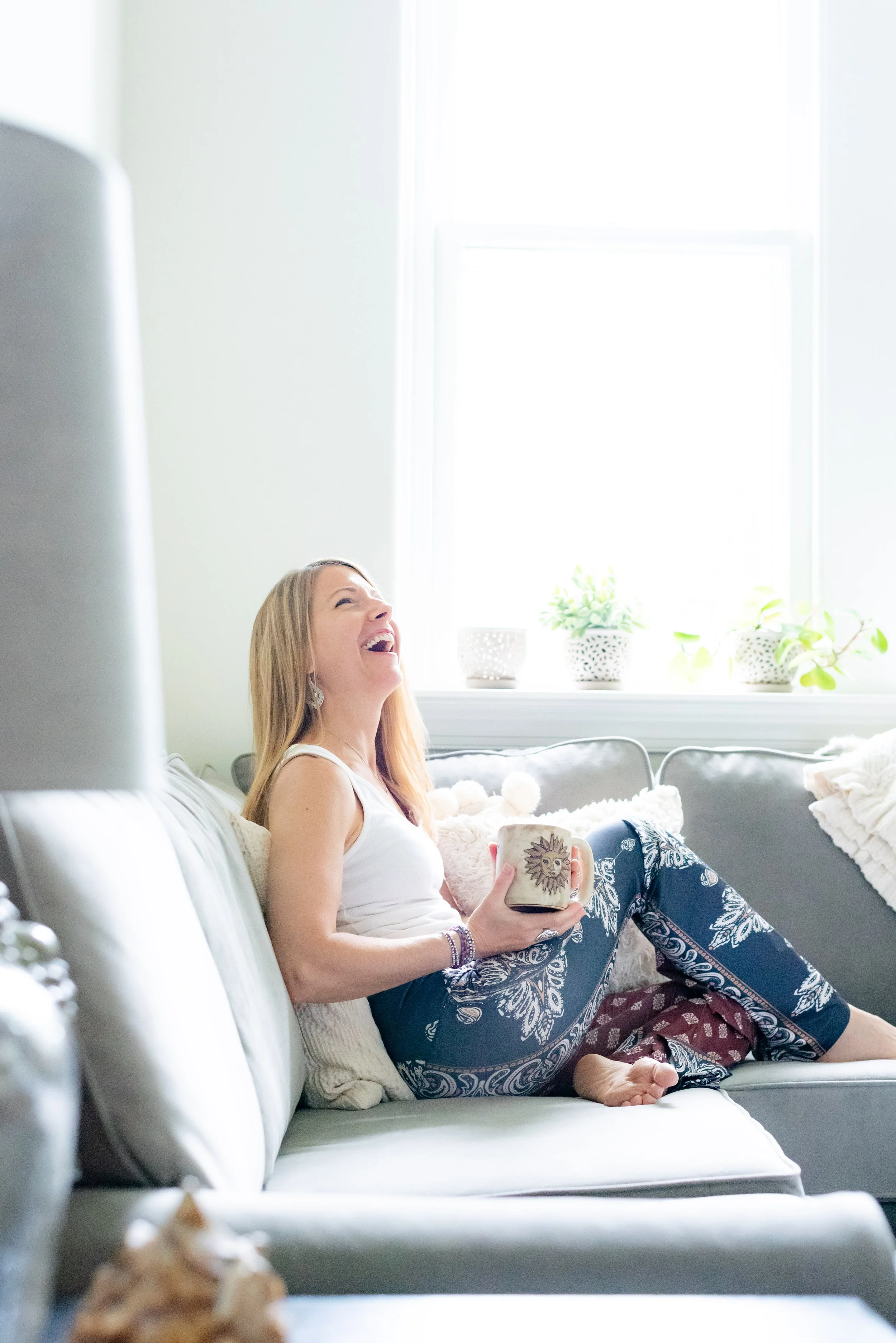 Parenting Astrologer, Tara Vogel wearing a white tank top and blue patterned pants, sitting on a light-colored sofa, laughing and holding a mug with a sun face design. She is in a bright room with a large window and potted plants on the windowsill.
