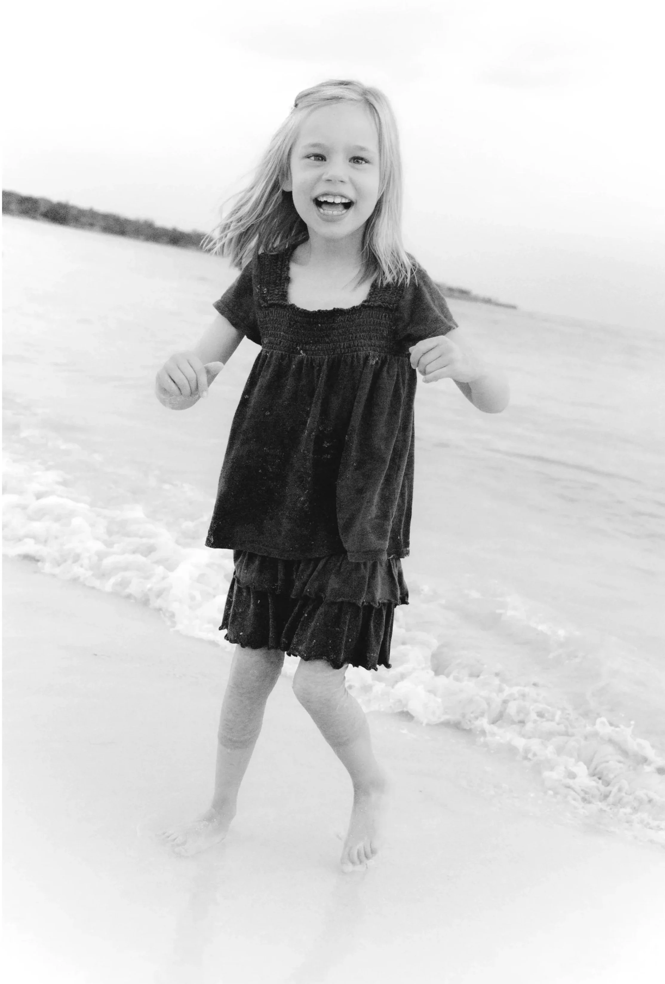 A young girl with long hair smiling and running along the shoreline at the beach, with the sea and sky in the background.
