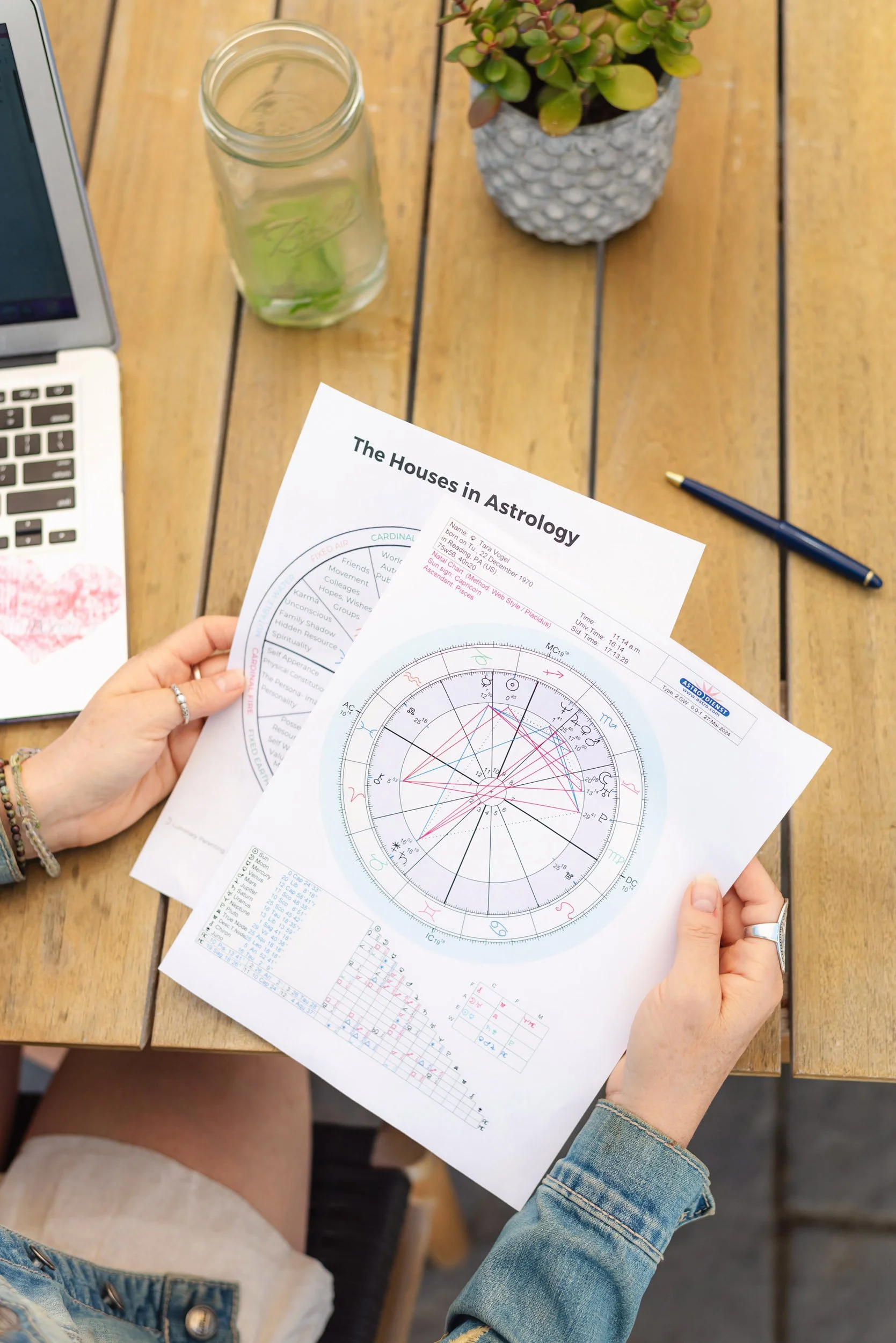 A woman holding printed astrology charts titled 'The Houses in Astrology' over a wooden table. The table has a laptop, a jar with a green plant, a blue pen, and a potted succulent.