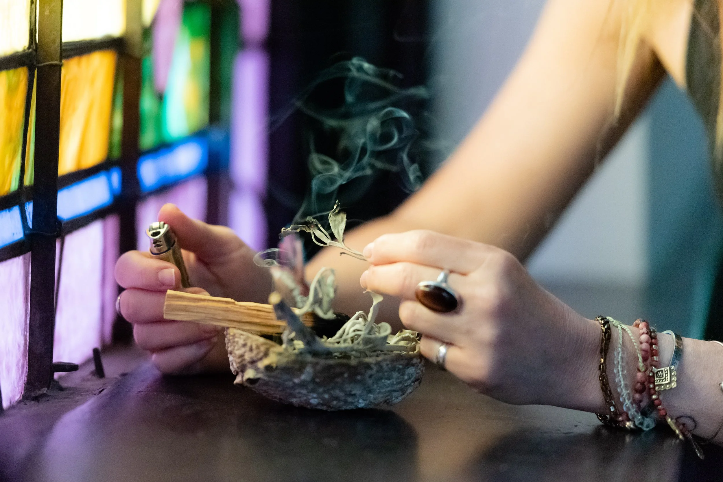 Close-up of a woman's hands holding a smoking herbal bundle and a lighter, near a colorful stained glass window.