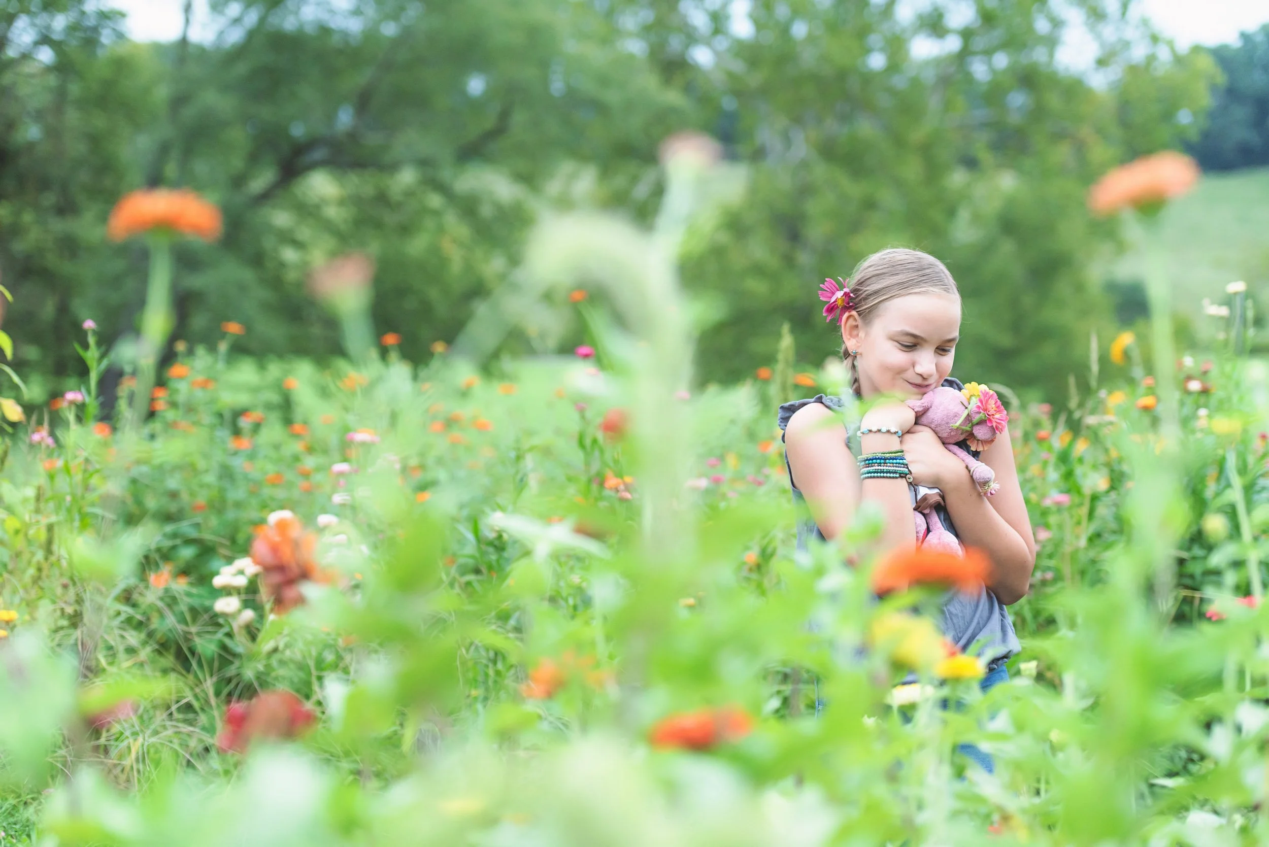 A girl hugging a stuffed animal in a field of colorful wildflowers with green trees in the background.