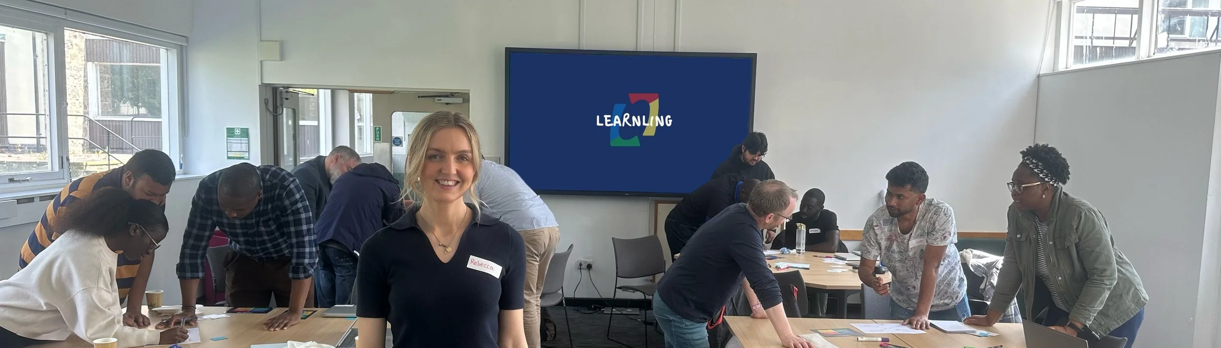 Group of diverse people gathered in a bright classroom or meeting room, engaged in a collaborative activity, with a smiling woman wearing a name tag in the foreground and a digital display with the word 'LEARNING' in the background.