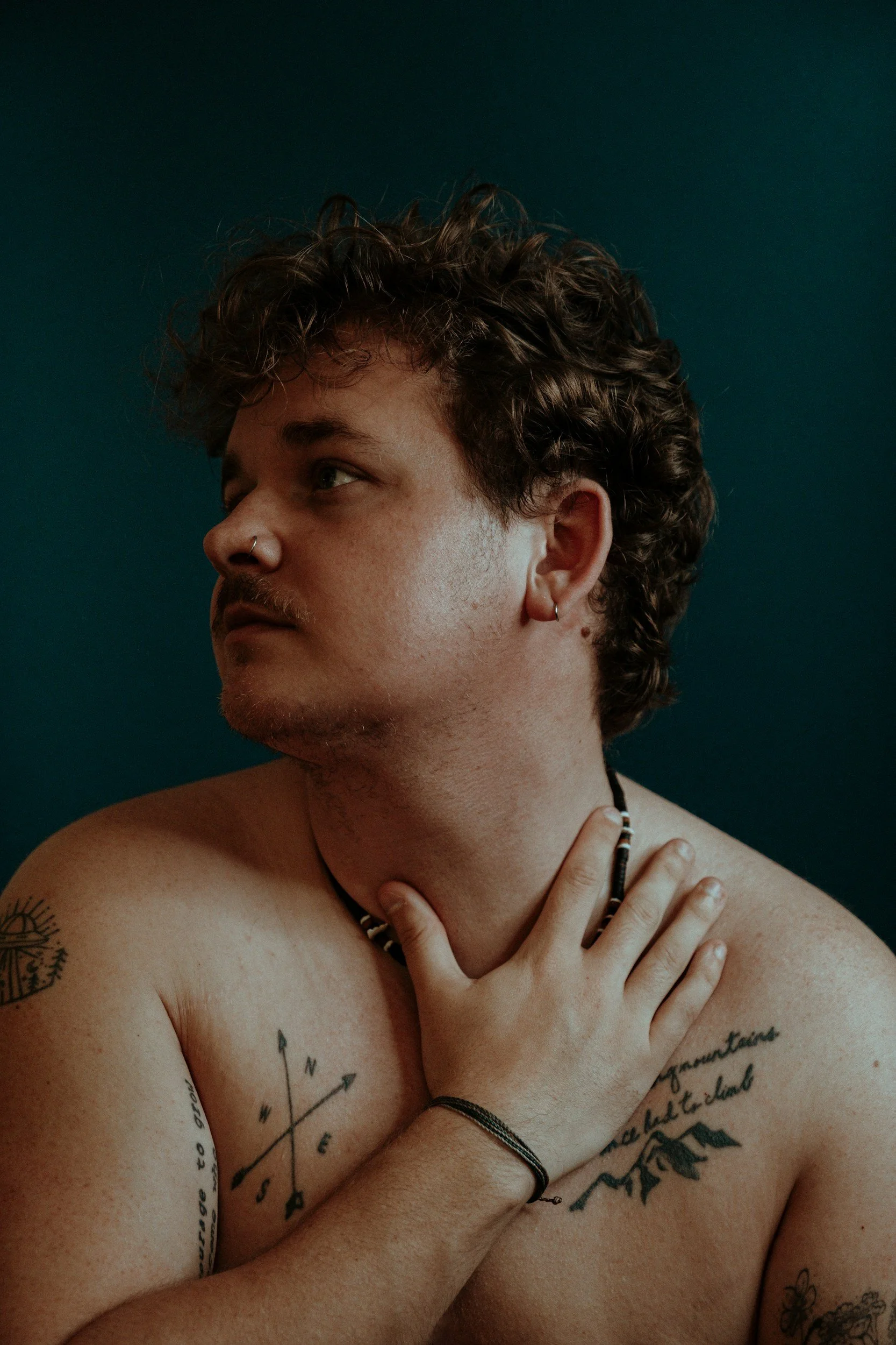 Close-up of a shirtless person with tattoos on their chest and arm, wearing earrings and necklaces, looking to the side with their hand resting on their chest, set against a dark teal background.