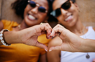 Two people making a heart shape with their hands, smiling, wearing sunglasses, outdoors.
