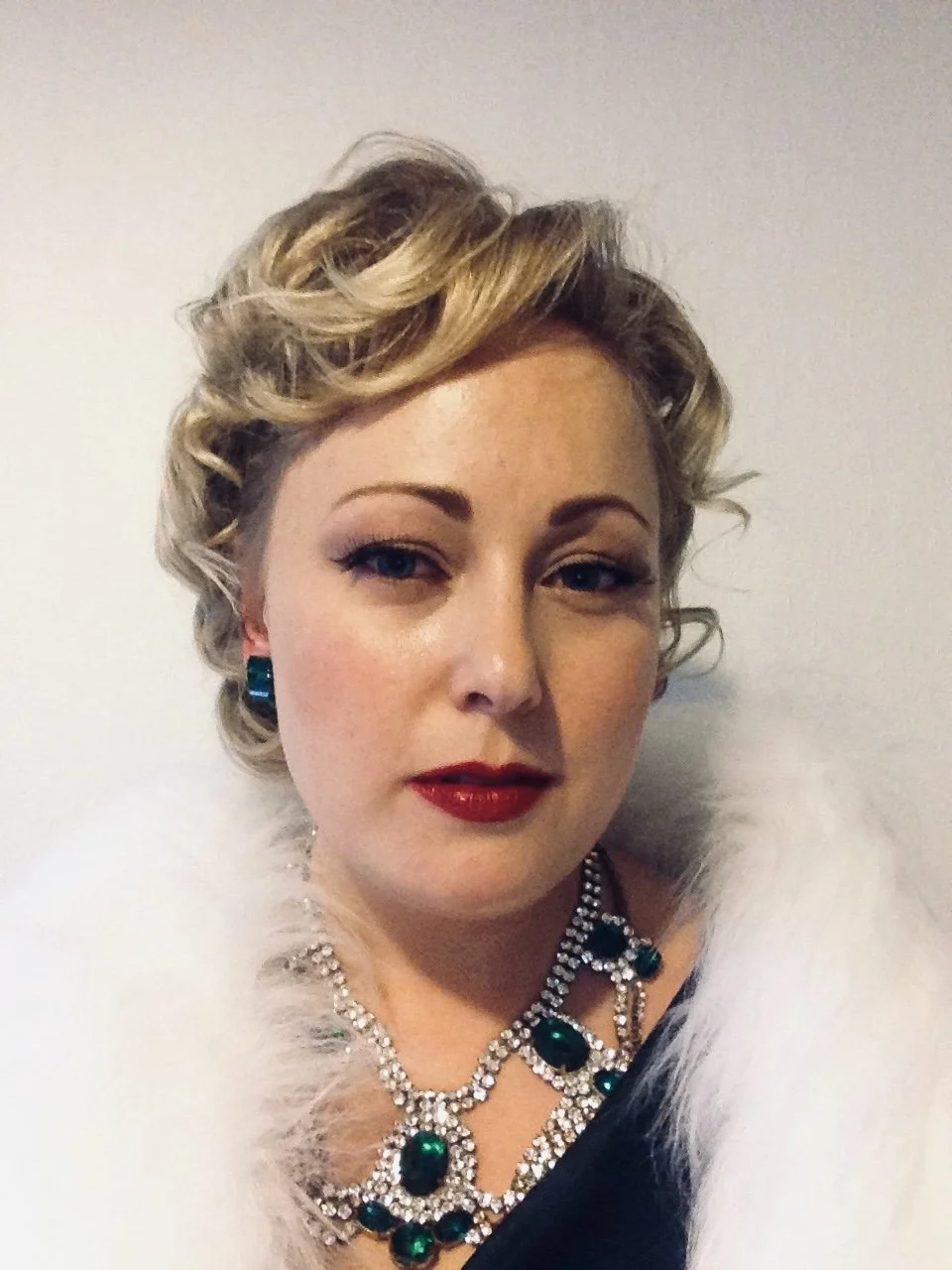 Kvinde med klassisk burlesque-stil, rød læbestift og glamourøst smykke med grønne sten iført hvid fjerboa.