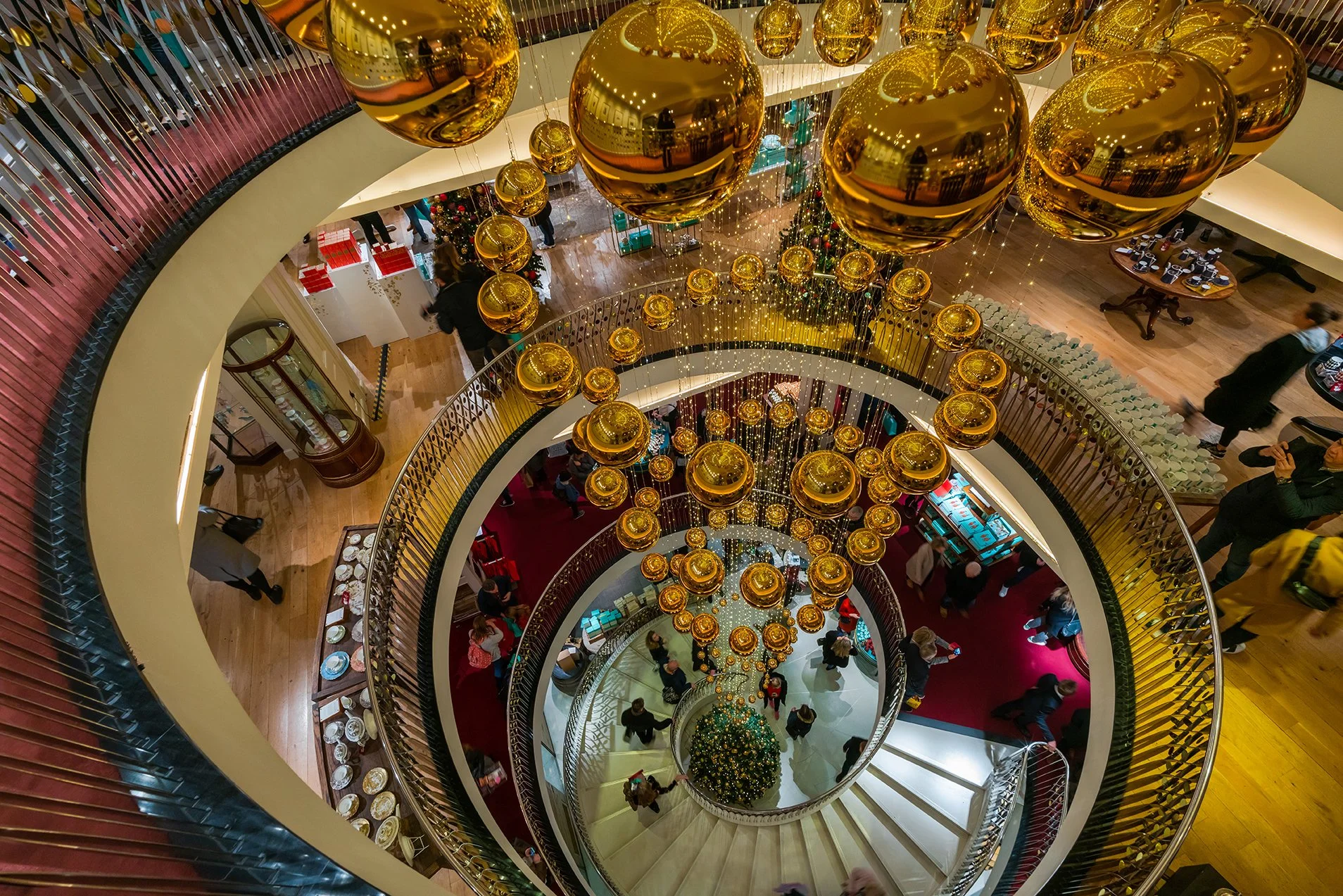 Decorated spiral staircase inside a shopping mall during Christmas with hanging gold ornaments and a Christmas tree at the bottom, shoppers walking around, and tables set for dining.
