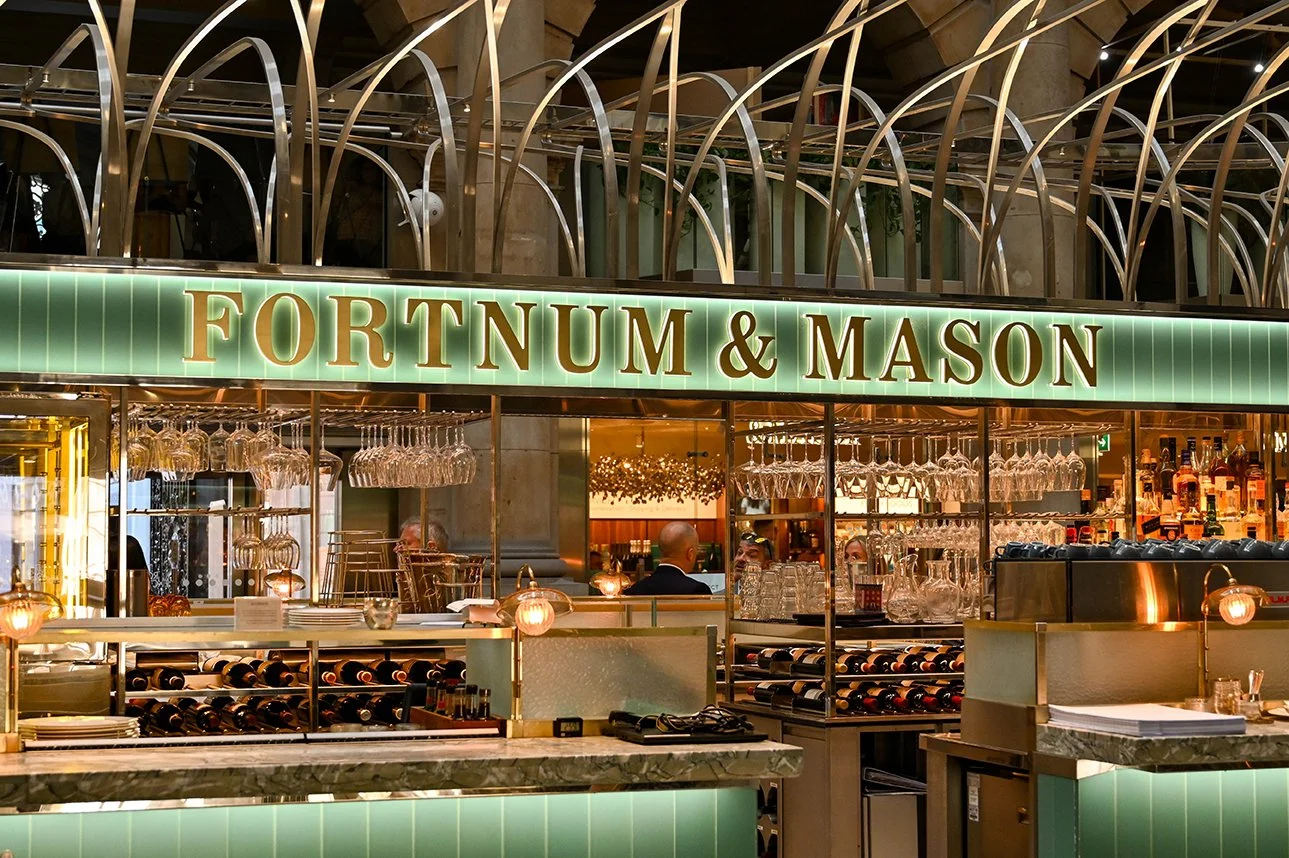 Close-up view of a gold-colored door handle on a textured glass door in a bright, modern interior with seating and plants in the background.