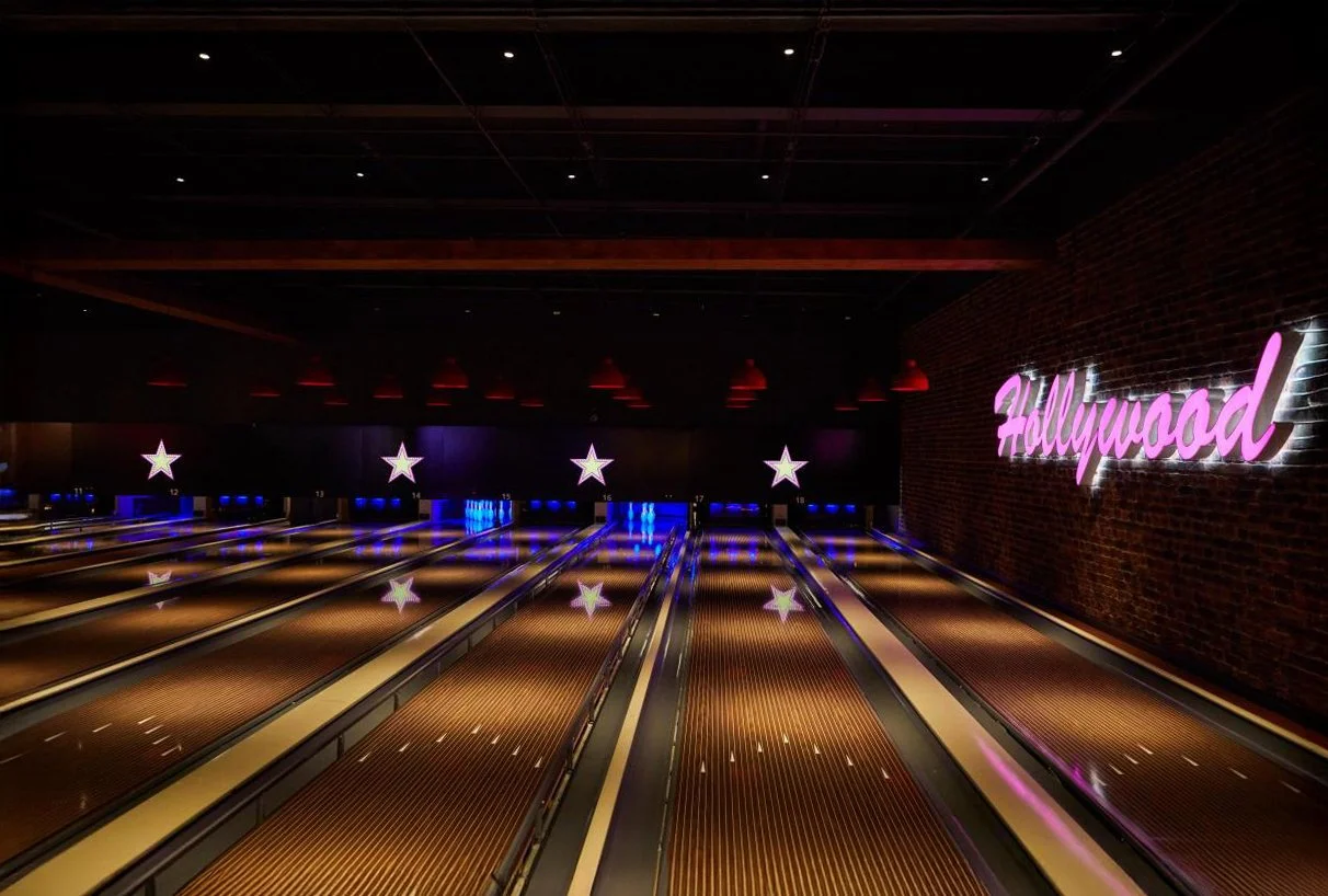 Empty bowling alley with multiple lanes, illuminated star decorations, and a neon pink Hollywood sign on a brick wall