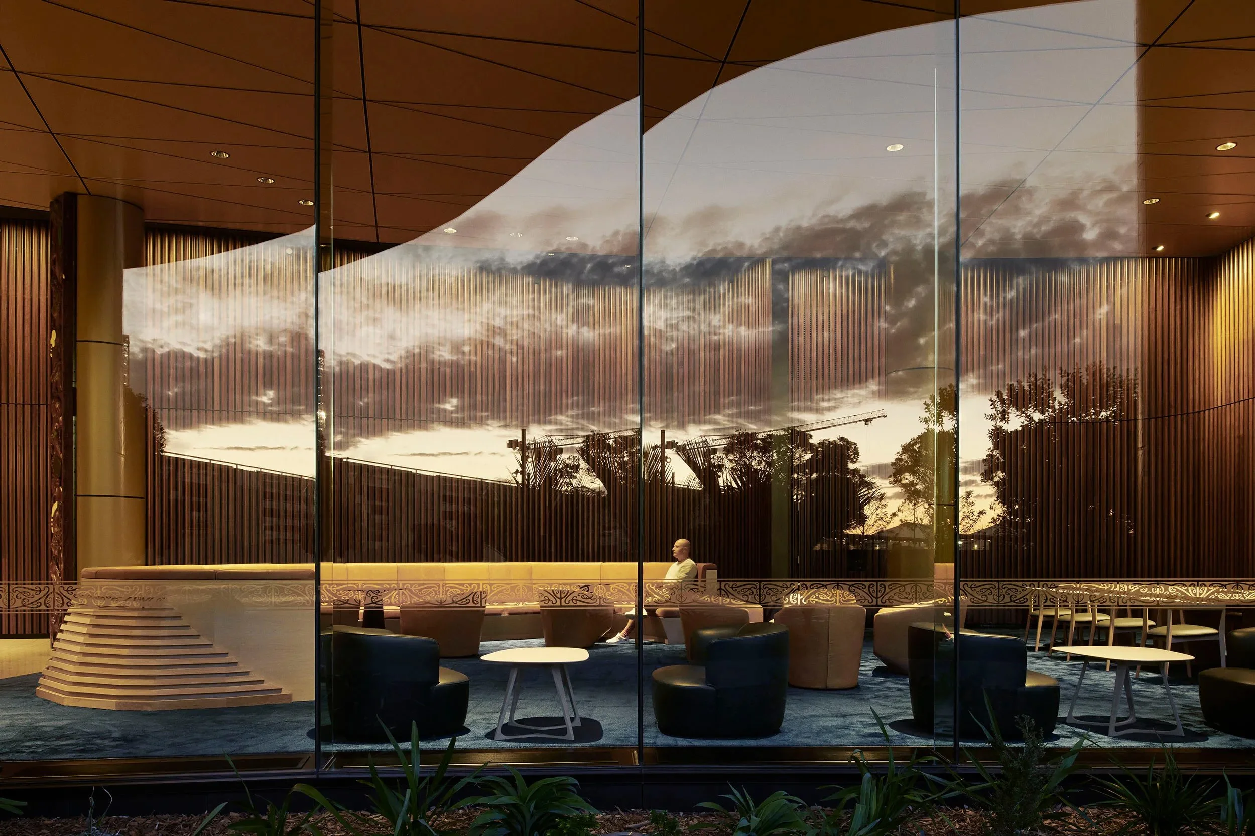 Interior of a modern building with large glass windows reflecting a sunset sky, and an elegant seating area with black and beige chairs, small tables, and a woman sitting near the window.