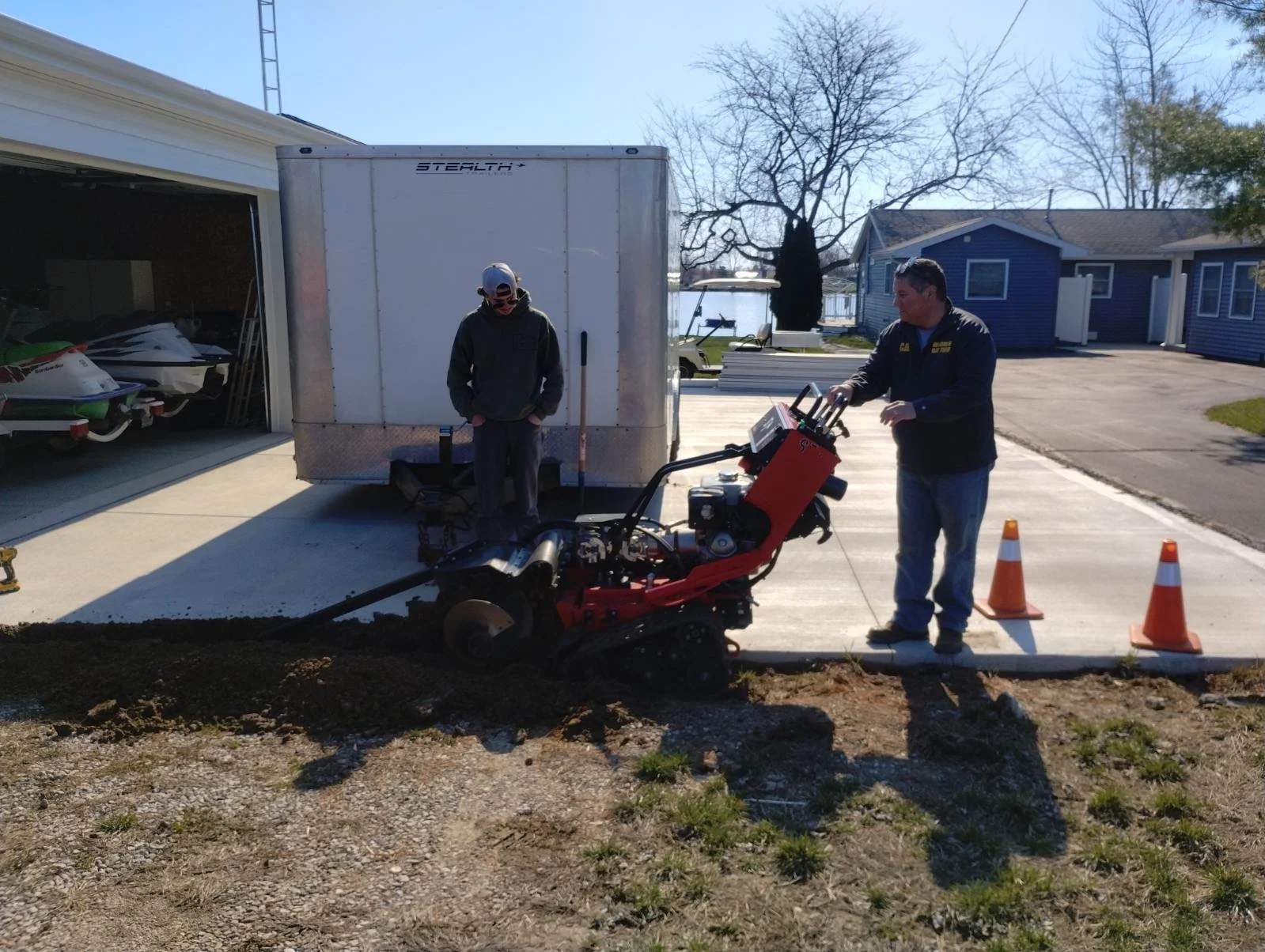 Two men working on a concrete driveway. One man is operating a small power trowel, and the other man is standing next to a trailer. There are orange traffic cones marking the area, and residential houses are visible in the background.