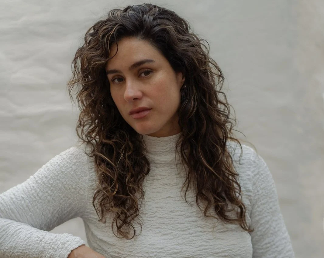 A man with curly dark hair, glasses, and a beard, wearing a dark green sweater, looking thoughtful against a neutral background with a plant in the background.