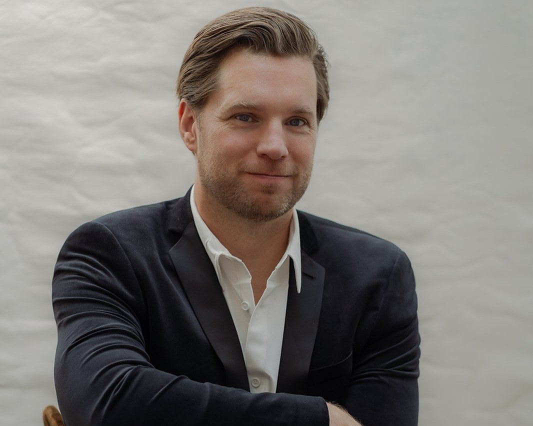 Smiling woman with curly hair, wearing glasses, hoop earrings, and layered necklaces, in an indoor setting.