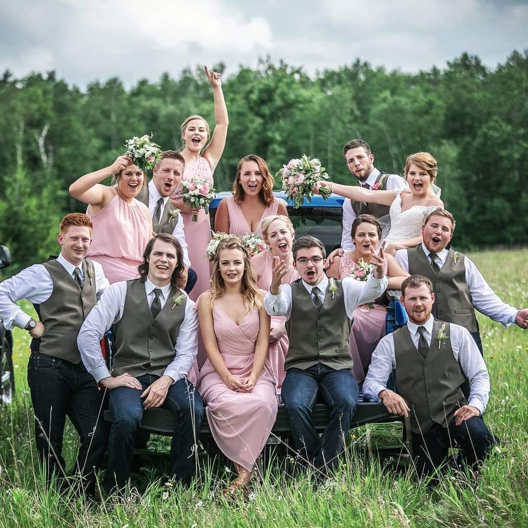 A wedding party posing outdoors in a field with trees in the background. The group includes bridesmaids in pink dresses, groomsmen in vests, and a bride in a white dress. They are sitting and standing on a vehicle, holding bouquets and cheering.