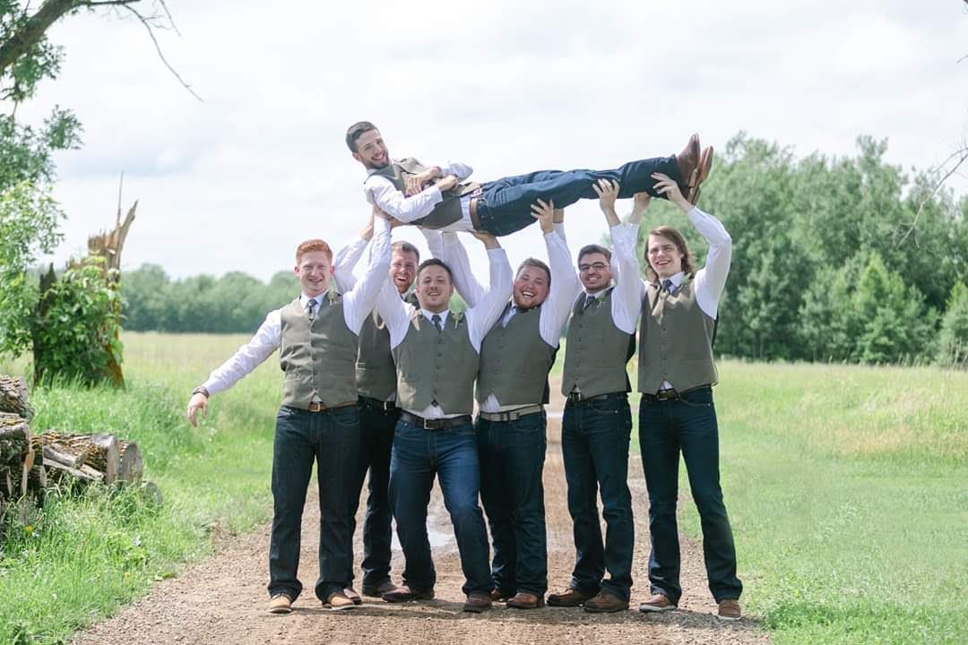 Group of groomsmen in vests and ties lifting a groom outdoors on a rural path.