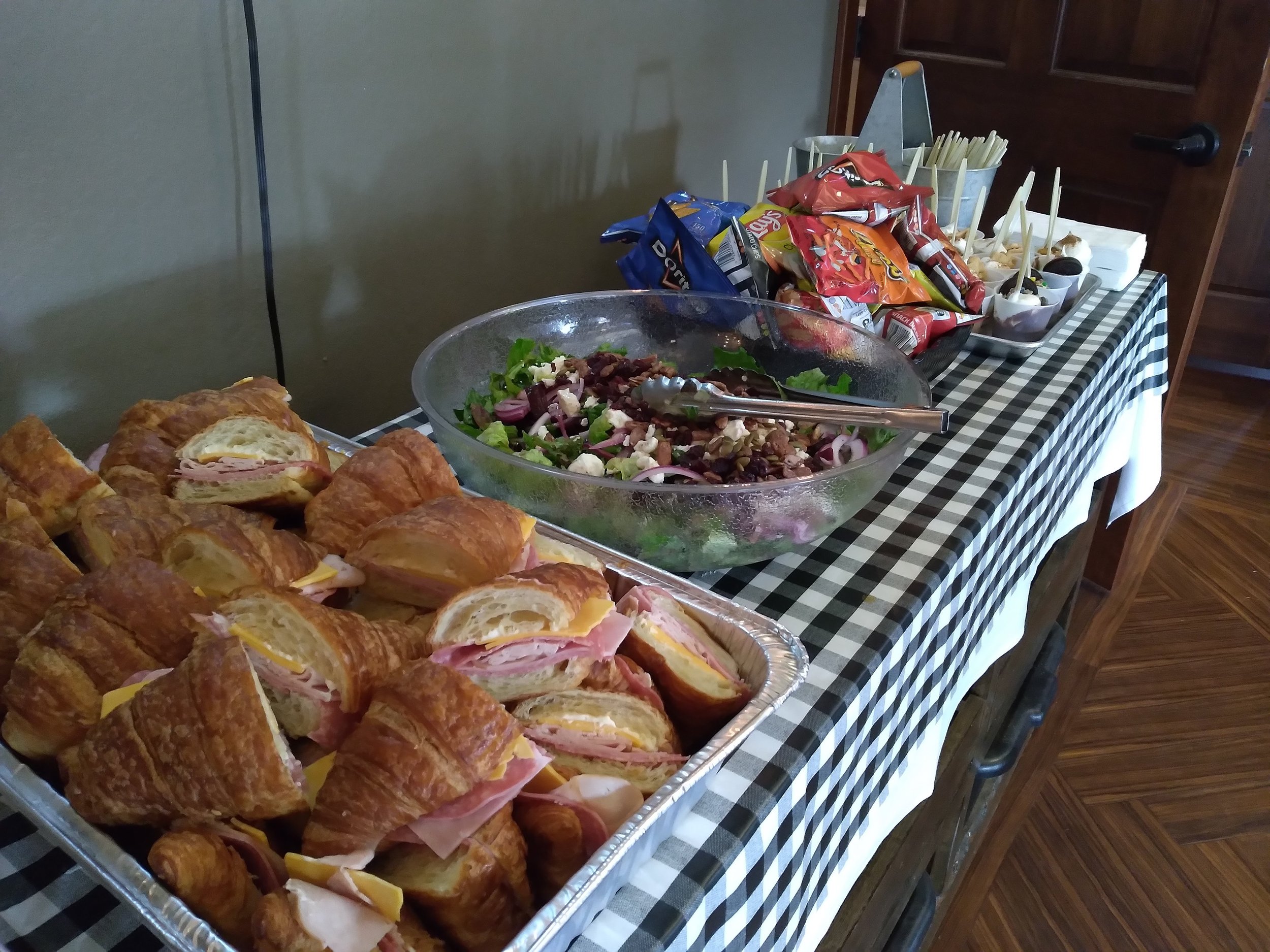 Buffet table at Hunts Point Gun Club for an event with a variety of croissant sandwiches, salad in a bowl, and various bags of chips on a checkered tablecloth.