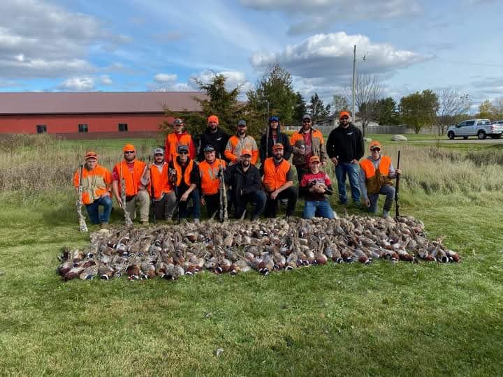 Group of hunters wearing orange vests posing with pheasants on grass in front of Hunts Point Gun Club.