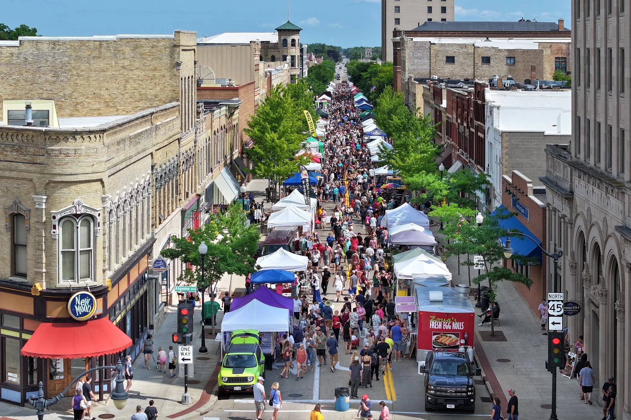 A crowded street market with vendor tents lined along both sides, bustling with people. Historic brick buildings flank the street, and a mix of colorful canopies and pedestrian activity characterize the lively scene. Trees provide shade, and a visible street sign indicates "One Way."