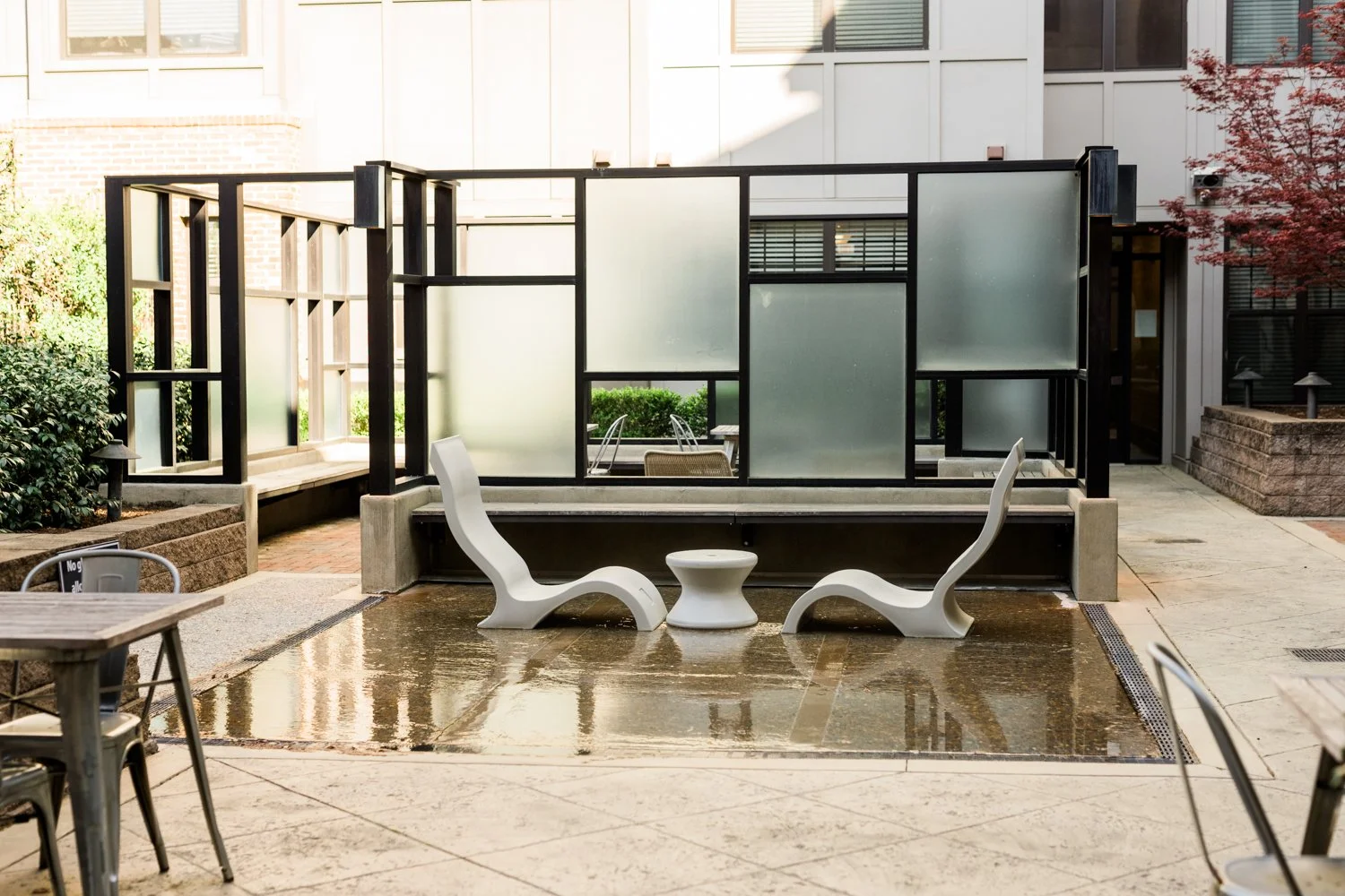 Modern outdoor patio area with two white lounge chairs and a small round table on a concrete surface, surrounded by frosted glass panels and greenery.