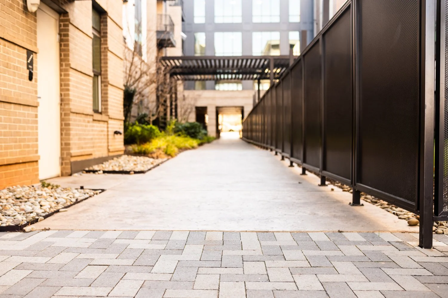 Modern urban walkway between buildings with brick walls and metal fence, lined with decorative rocks and plants.