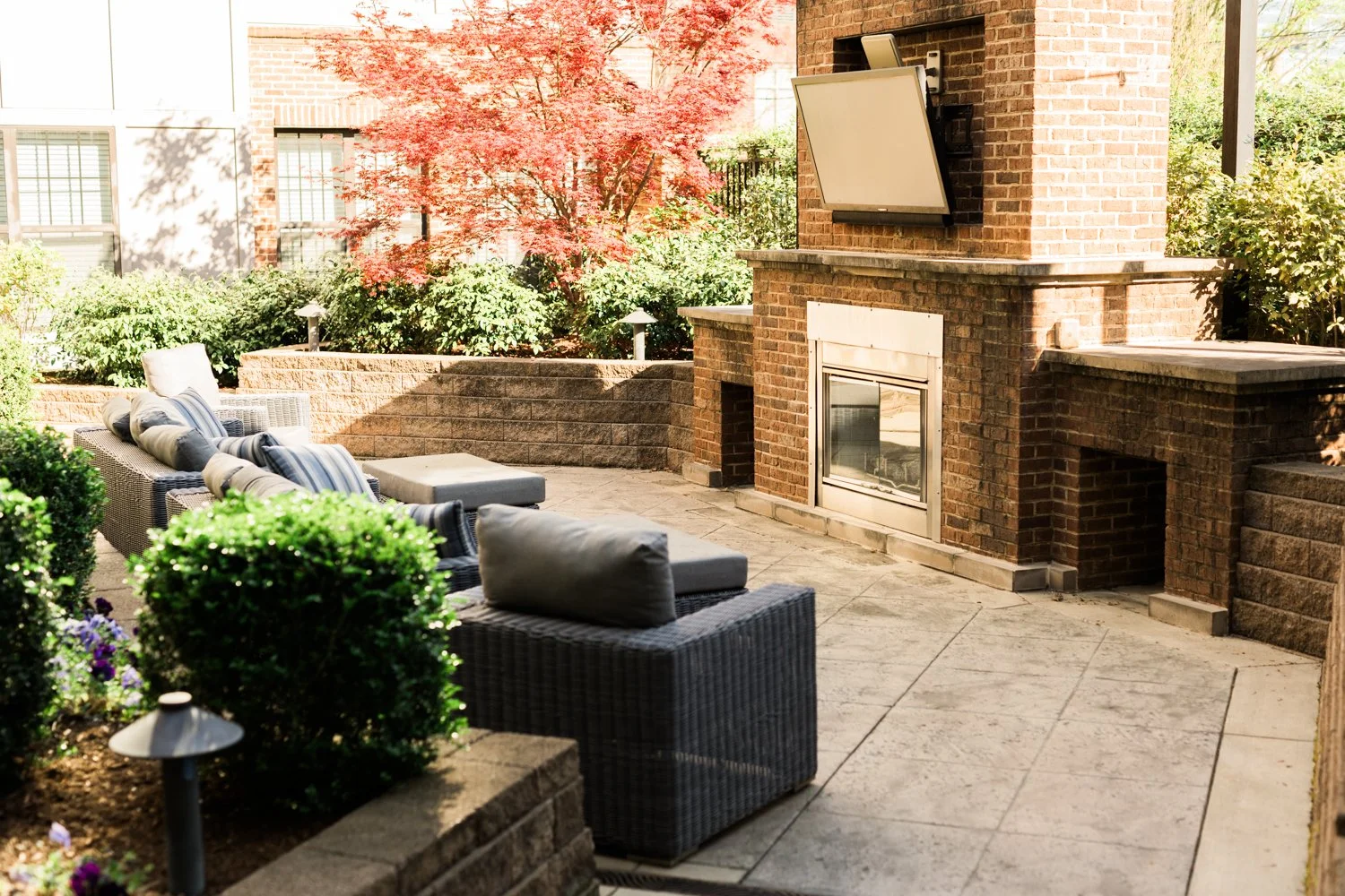 Outdoor patio with wicker furniture, fireplace, mounted TV, and lush greenery including a Japanese maple tree.
