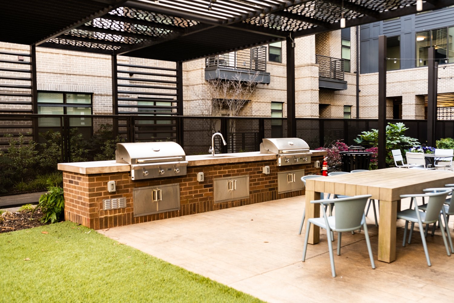 Outdoor kitchen area with barbecue grills, sink, table, and chairs under a pergola, surrounded by brick walls and landscaping.