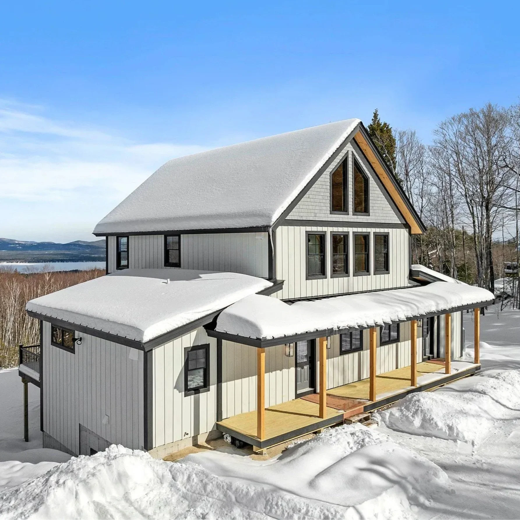 Snow-covered house with a porch, surrounded by snow and trees under a clear blue sky.
