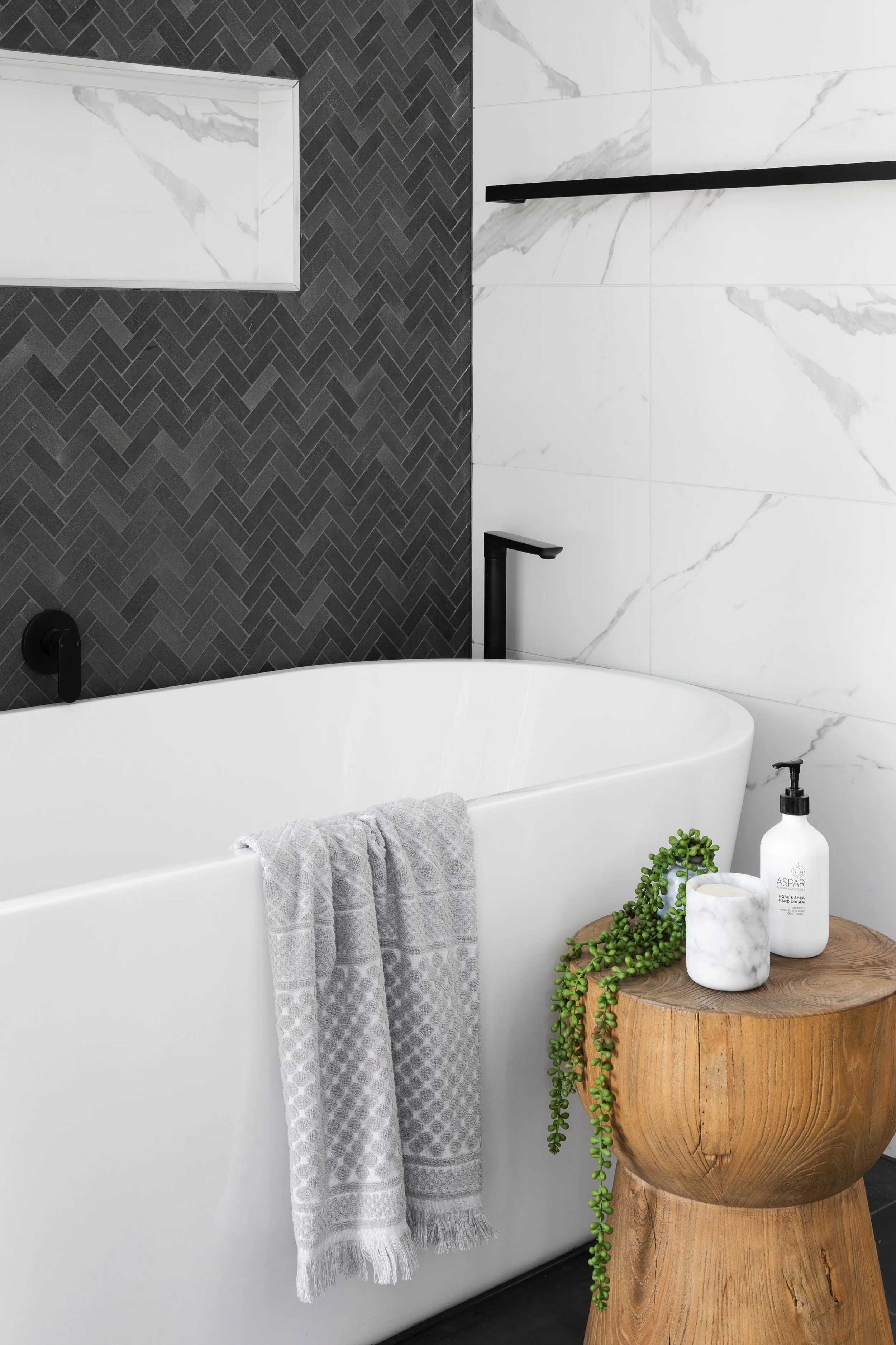Modern bathroom with freestanding white bathtub, black faucets, dark herringbone tile wall, white marble accent tiles, gray patterned towel, wooden stool with plant, candle, and soap dispenser.
