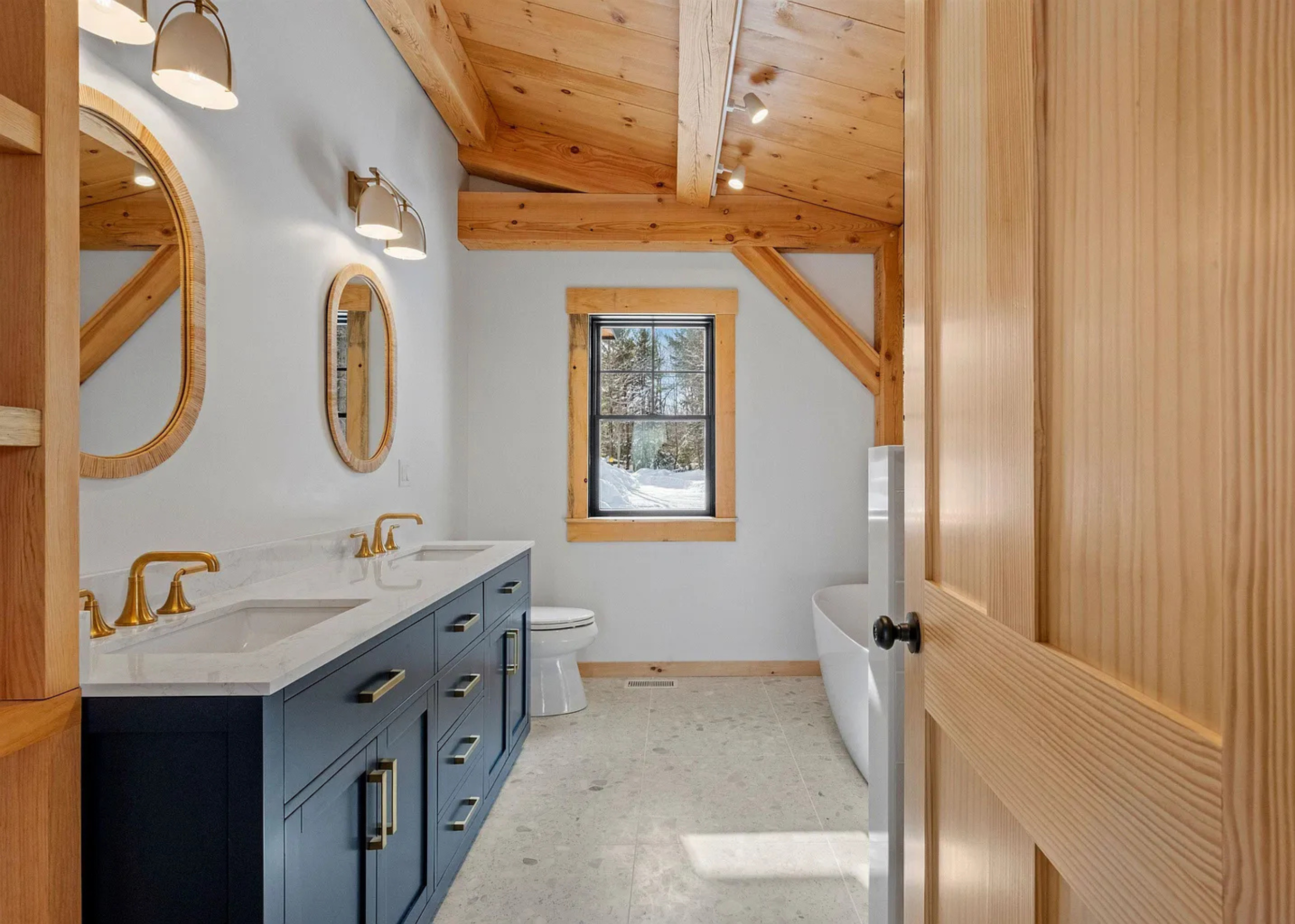 Modern rustic bathroom with double sink vanity, gold fixtures, white countertop, oval mirrors, tub, and wooden beams on ceiling.