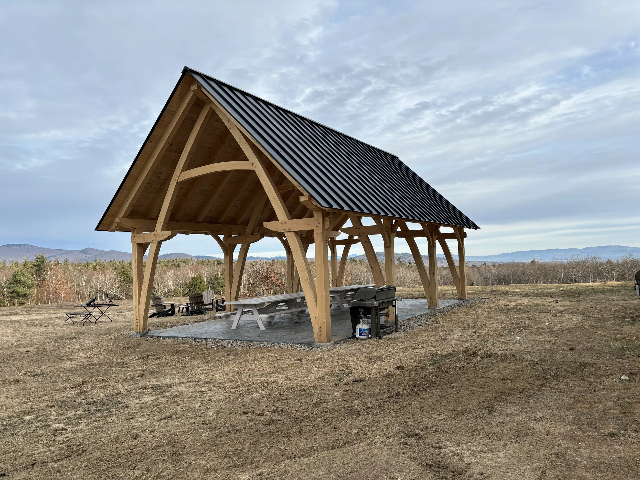 Timber-framed pavilion with a black metal roof in a field, surrounded by wooden chairs and distant trees.