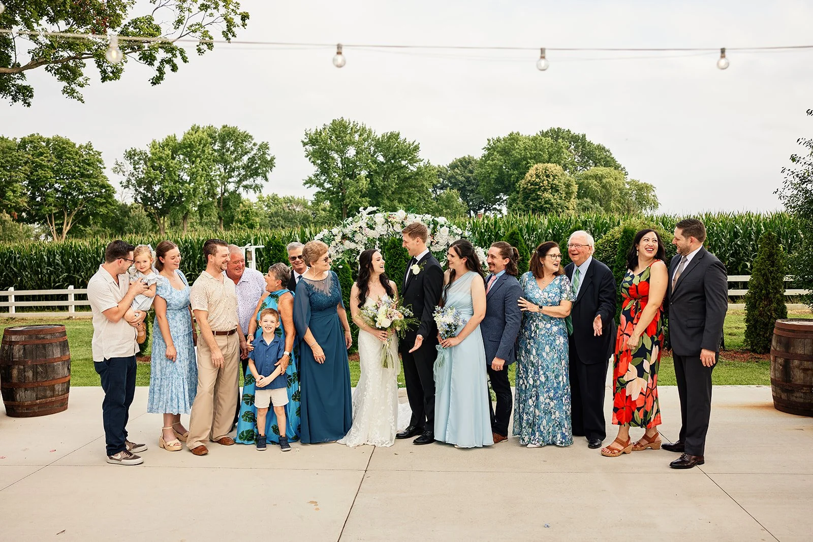 A group of people dressed in formal attire standing outdoors at a wedding, with the bride and groom at the center, surrounded by family and friends, in front of a floral arch and green trees. Bridal party, wedding day.