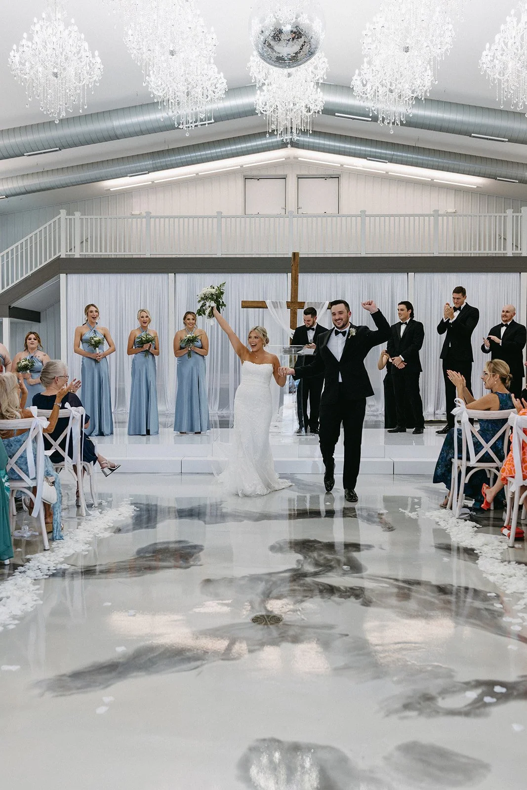 Bride and groom walking down the aisle after wedding ceremony, with wedding party and guests clapping and celebrating in a decorated indoor venue with chandeliers and a cross in the background.