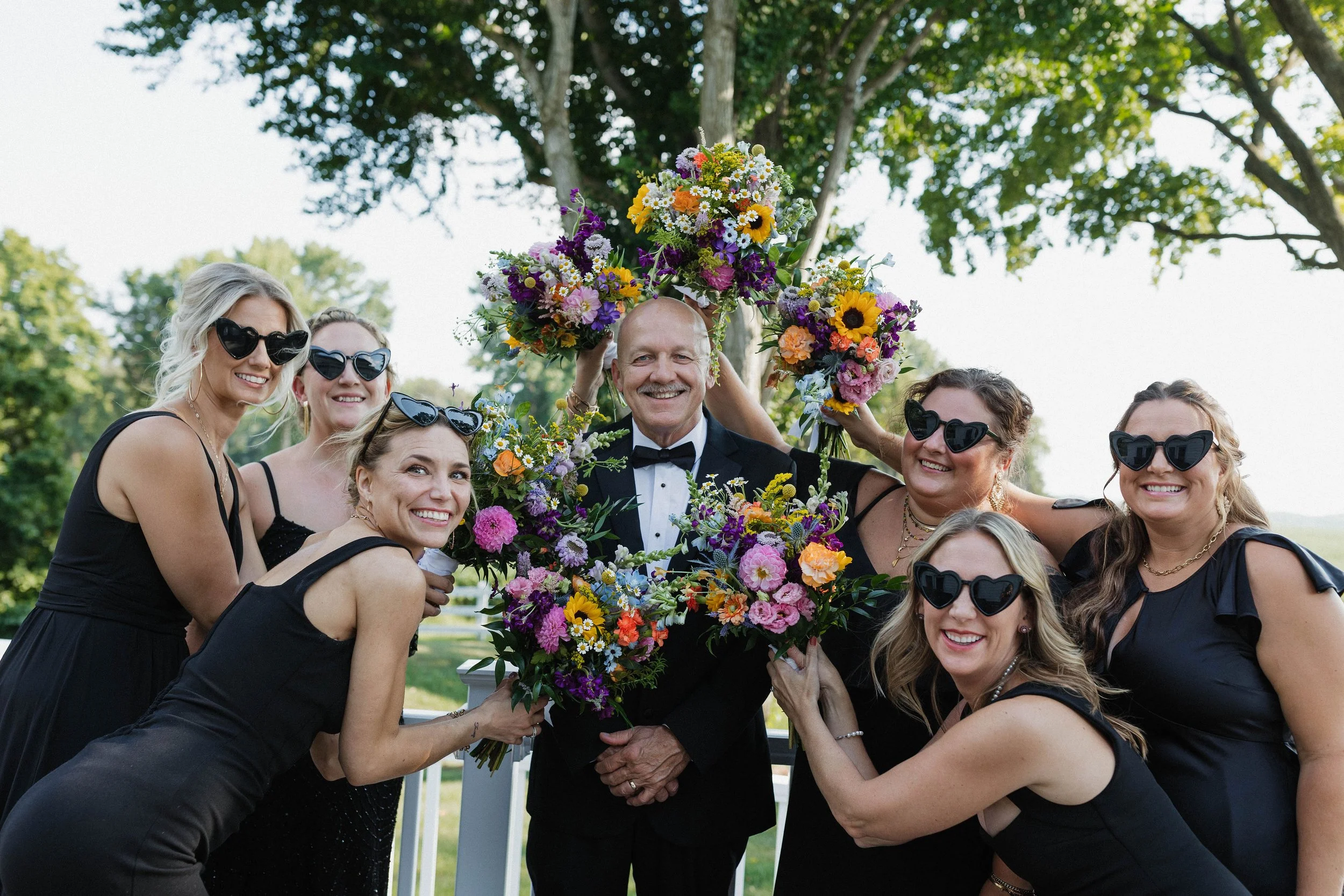 Group of women in black dresses and sunglasses holding colorful flower bouquets around a smiling man in a tuxedo, outdoors during daytime.