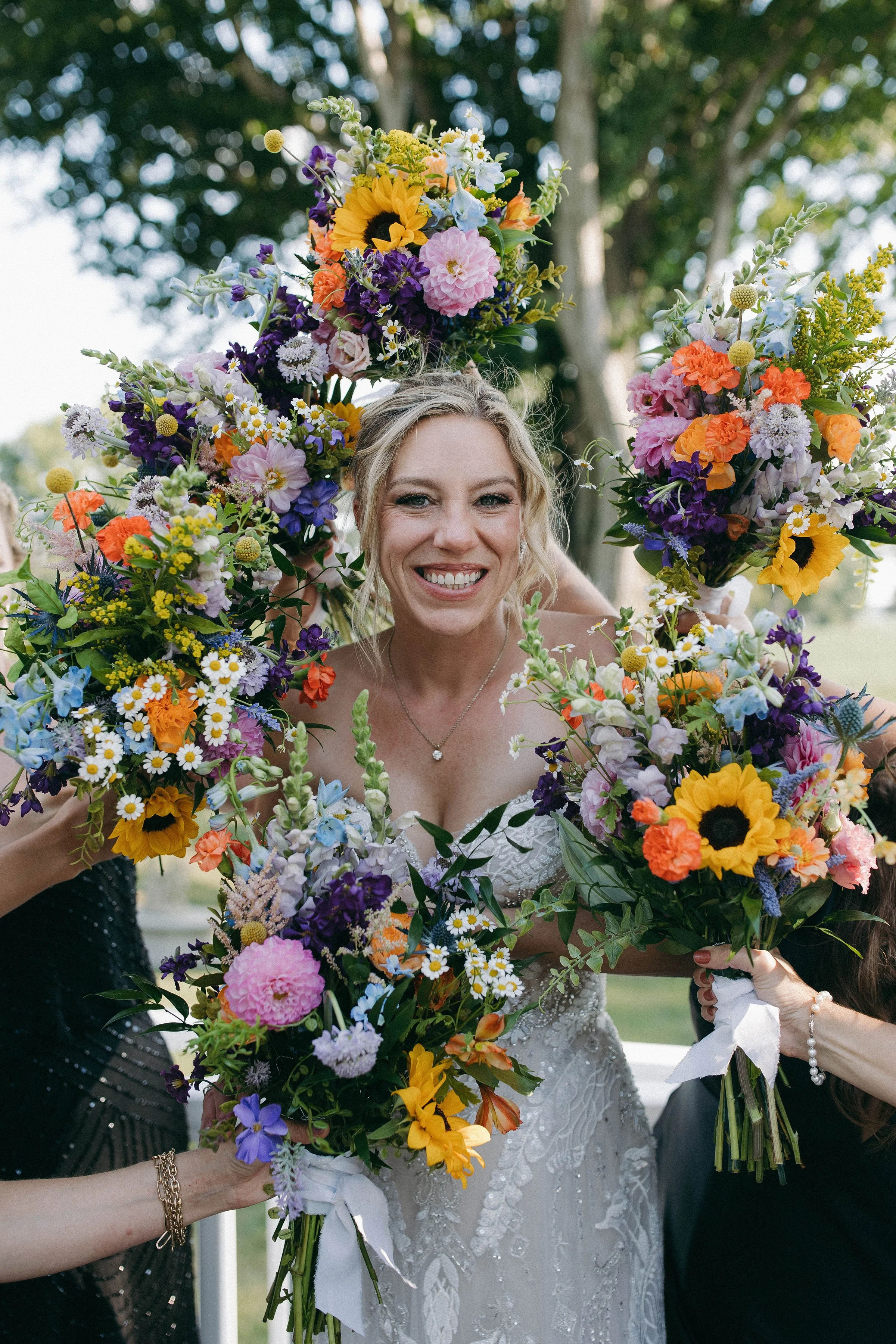 A smiling bride in a wedding dress holding a bouquet of colorful flowers, surrounded by friends holding similar bouquets at an outdoor celebration.