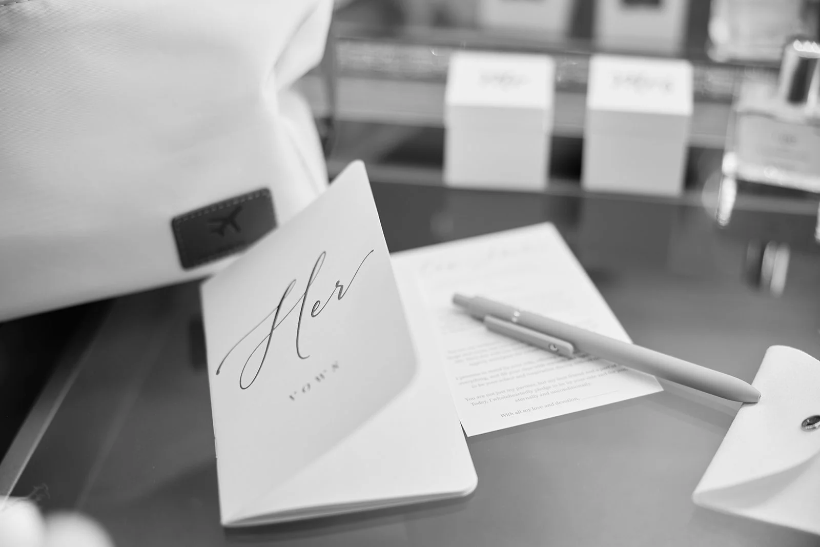 A table with a greeting card that says 'her' in elegant script, a pen, and some documents. In the background, there are boxes and a bottle.