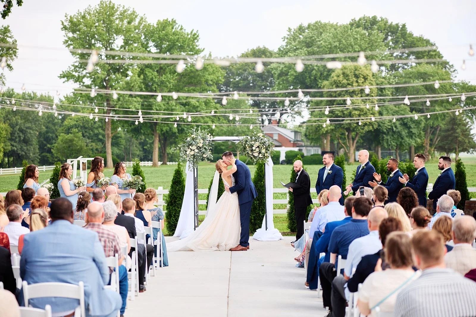 A wedding ceremony outdoors with a bride and groom kissing under an arch decorated with white flowers. Guests seated and standing on either side, with a view of trees and a house in the background, and string lights overhead.