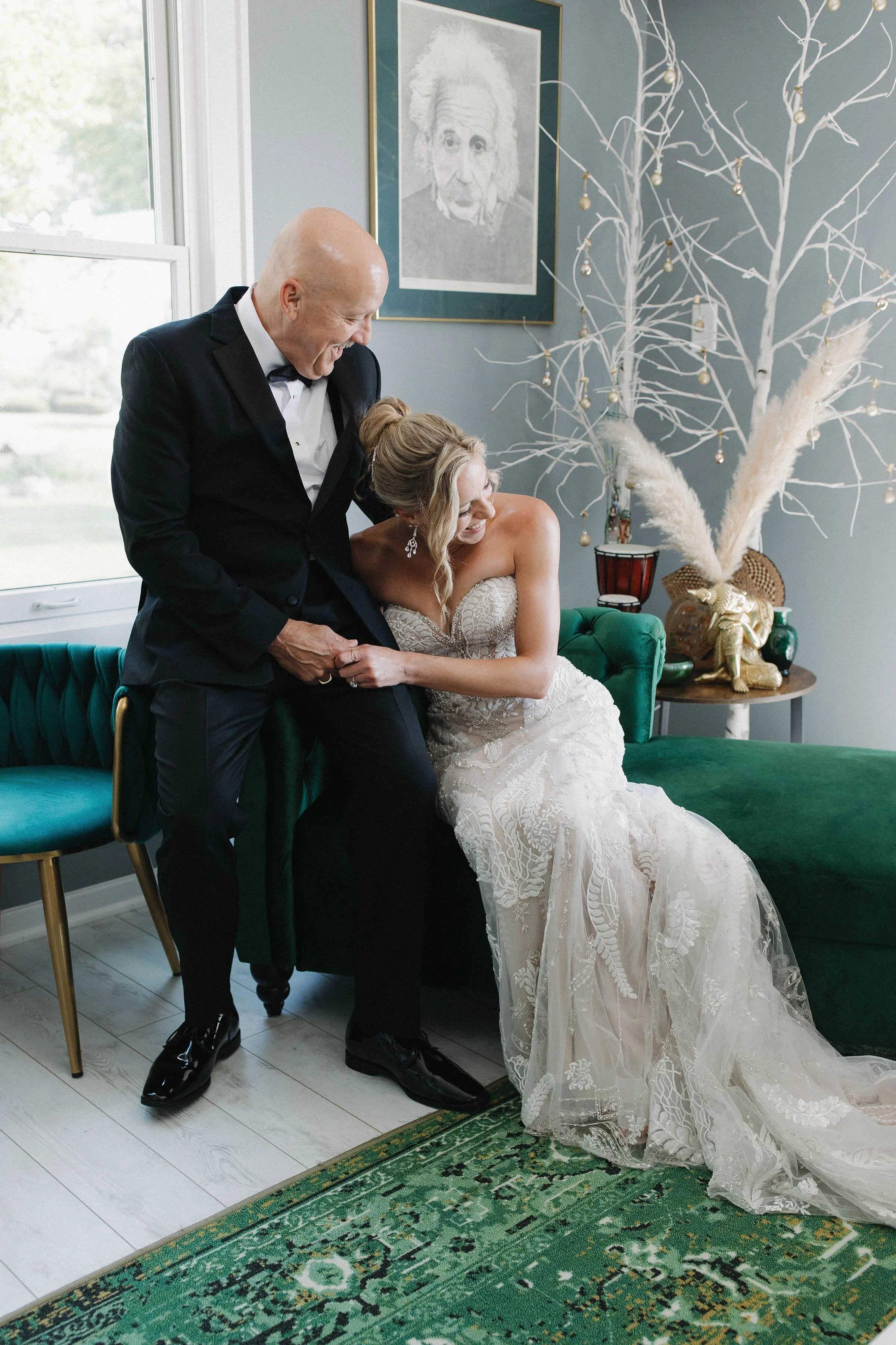 A bride in a white wedding dress sitting on a green velvet couch, being playful with an older man in a tuxedo, likely her father, in a cozy decorated room with artwork and a decorative tree in the background.