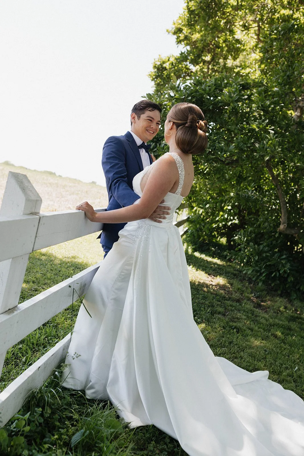 While leaning against a white fence, a groom in his navy blue suit smiles at his soon to be bride and holds her by the waist.