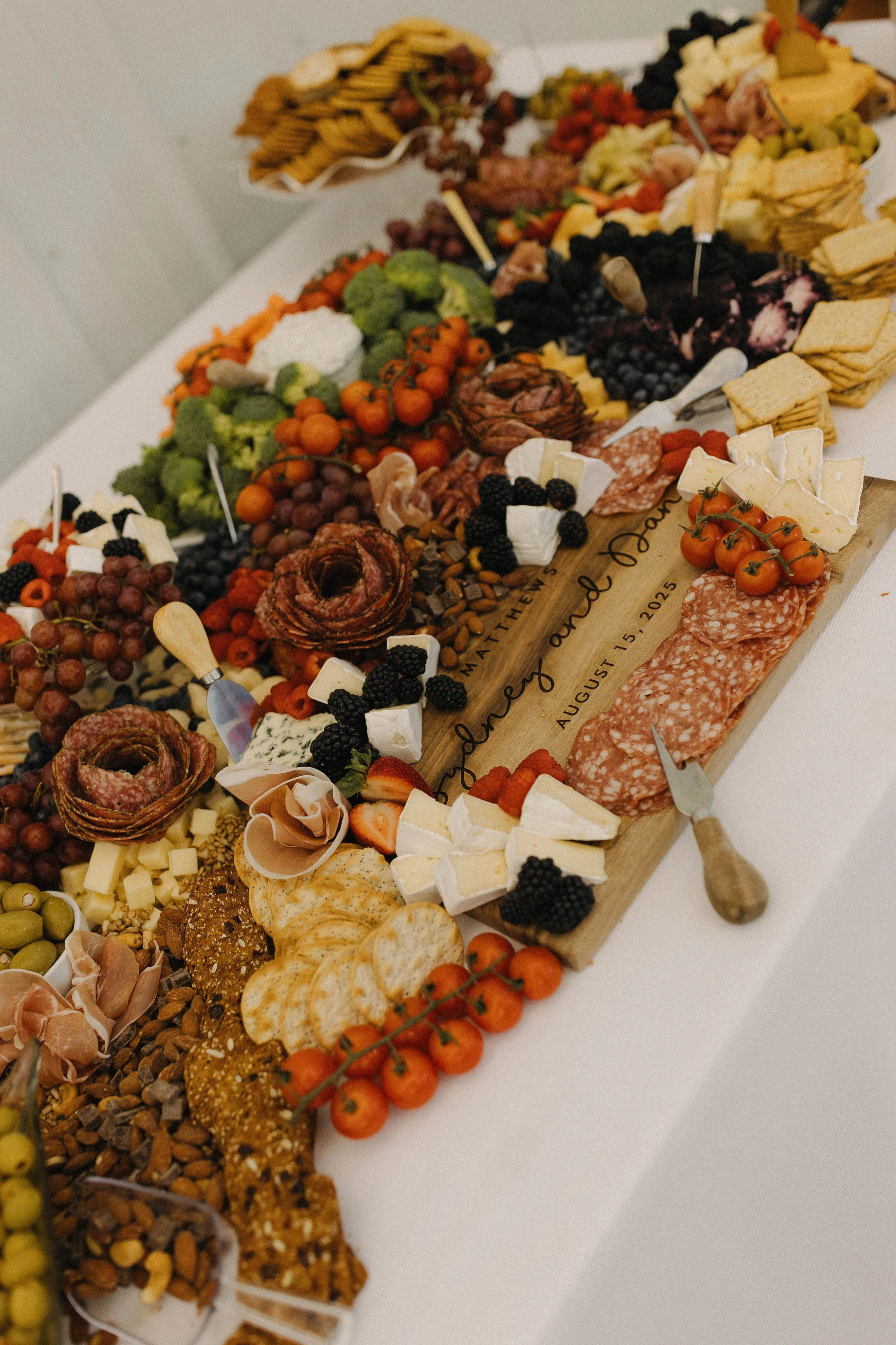 Charcuterie board with cheeses, meats, fruits, crackers, and vegetables on a wooden serving board, with some text engraved on it, set on a white table.