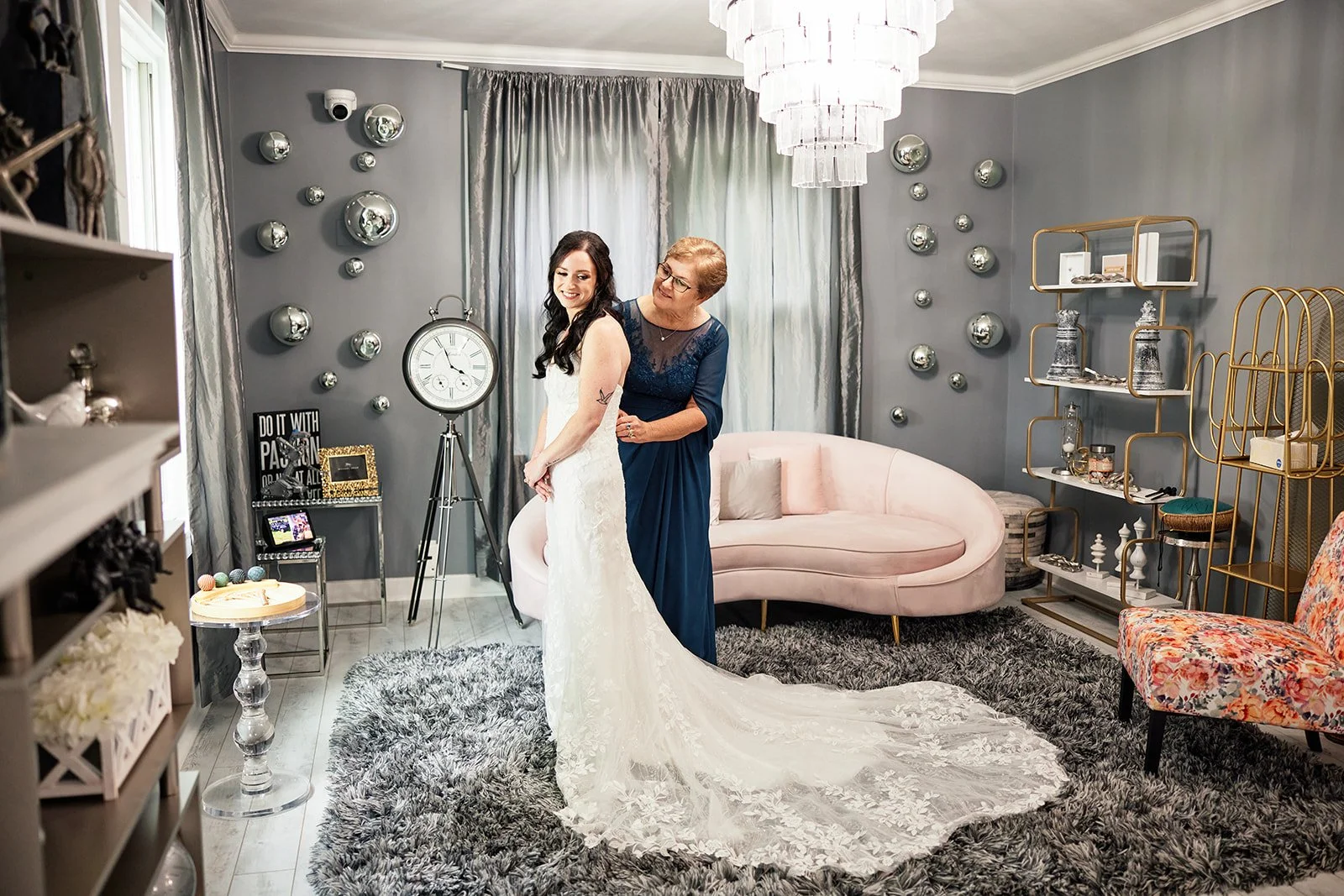 A woman in a white wedding dress is being helped by an older woman in a blue dress with her wedding gown in a decorated room with gray walls, silver wall art, a pink sofa, a gold shelf, and a shaggy rug.