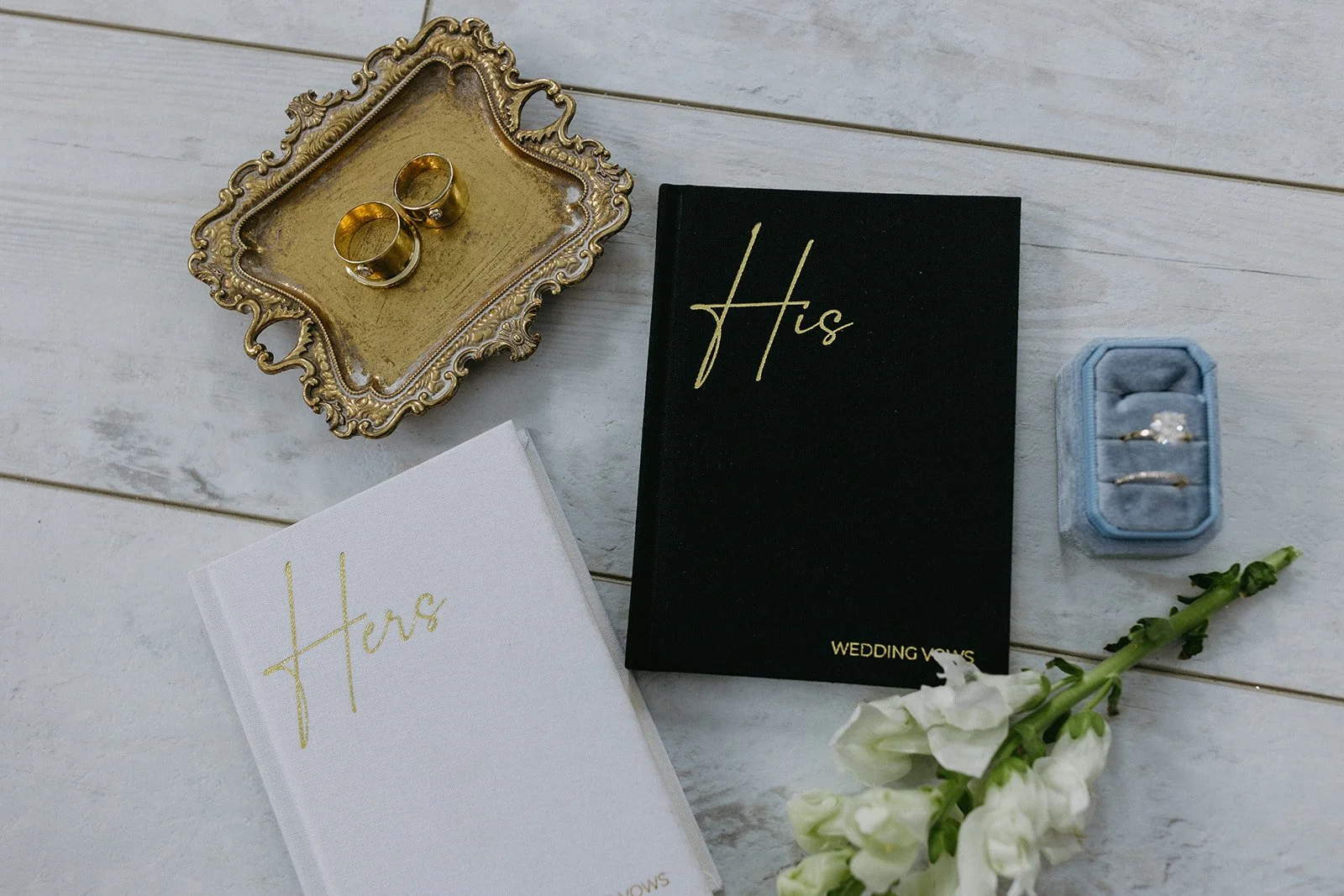 Wedding accessories including gold rings in a blue ring box, two wedding vow books labeled 'Hers' and 'His', a decorative tray, and white flowers arranged on a white wooden surface.