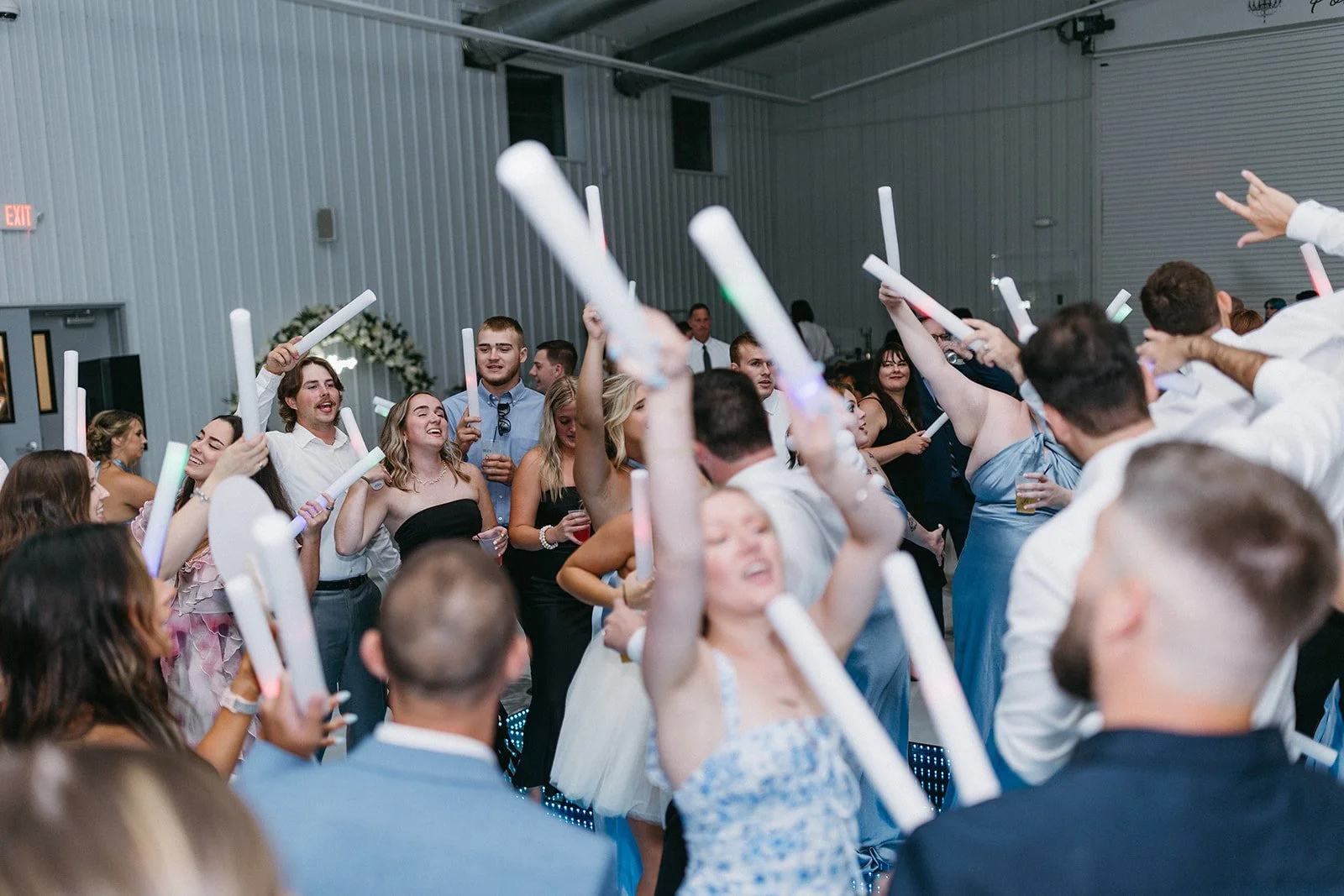 People dancing and celebrating with glow sticks at a party or wedding reception in a large indoor venue.