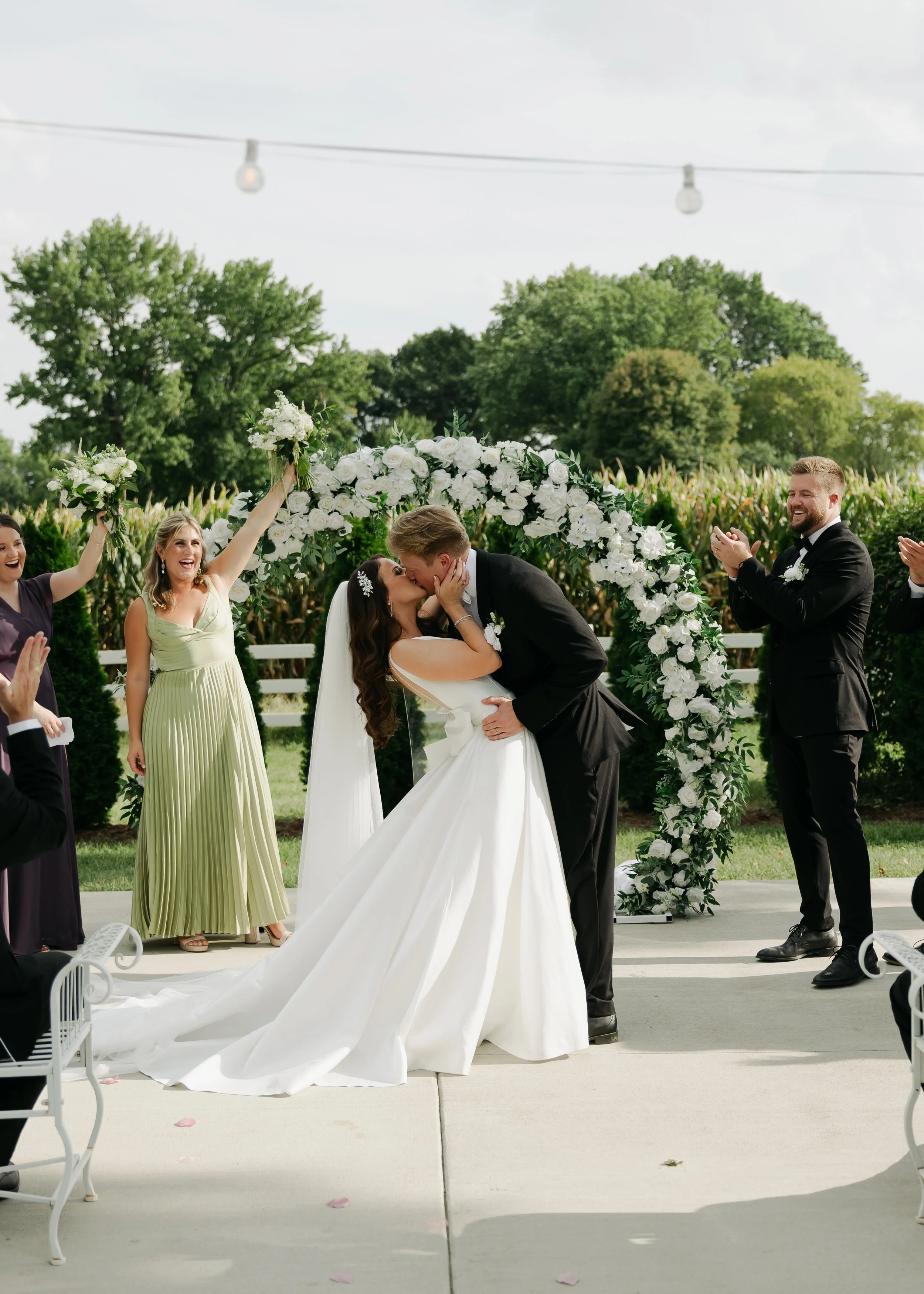 A bride and groom kissing under an arch of white flowers during an outdoor wedding ceremony at Pole Barn Chic in Central Illinois, Springfield IL.  surrounded by happy guests clapping.